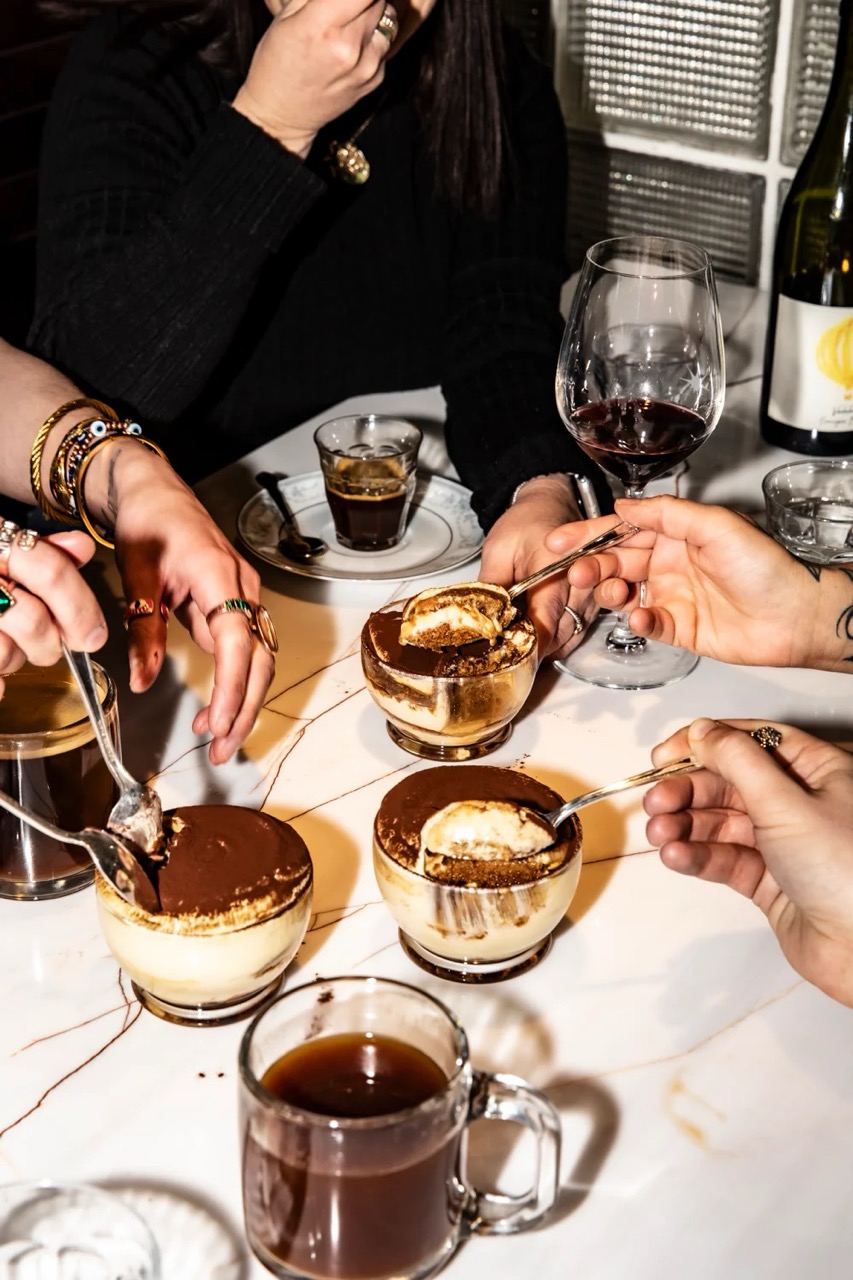 Three people sharing tiramisu desserts with spoons at a table, accompanied by coffee and a glass of red wine.
