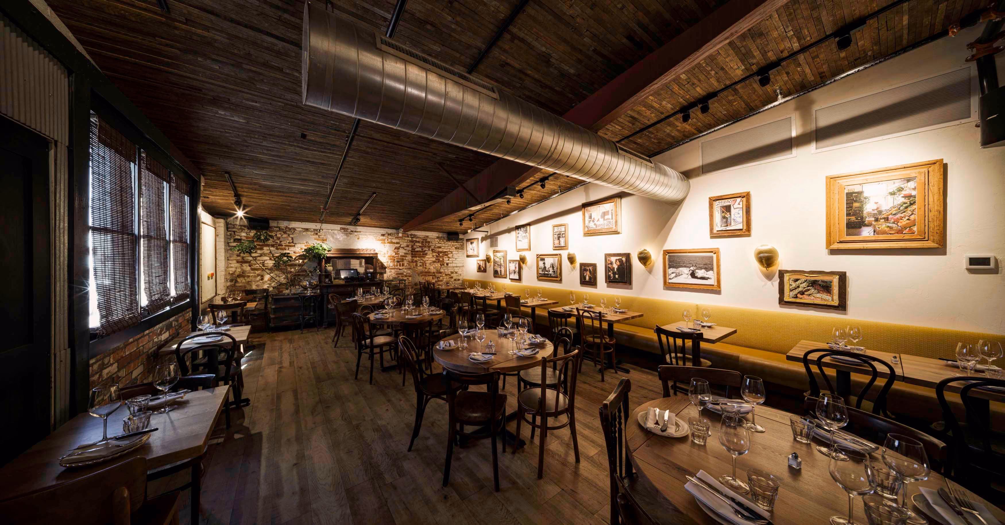 Cozy rustic restaurant interior with wooden floors, round and rectangular dining tables set with glassware, a yellow banquette along the wall, framed artworks, and exposed ductwork on a wooden ceiling.