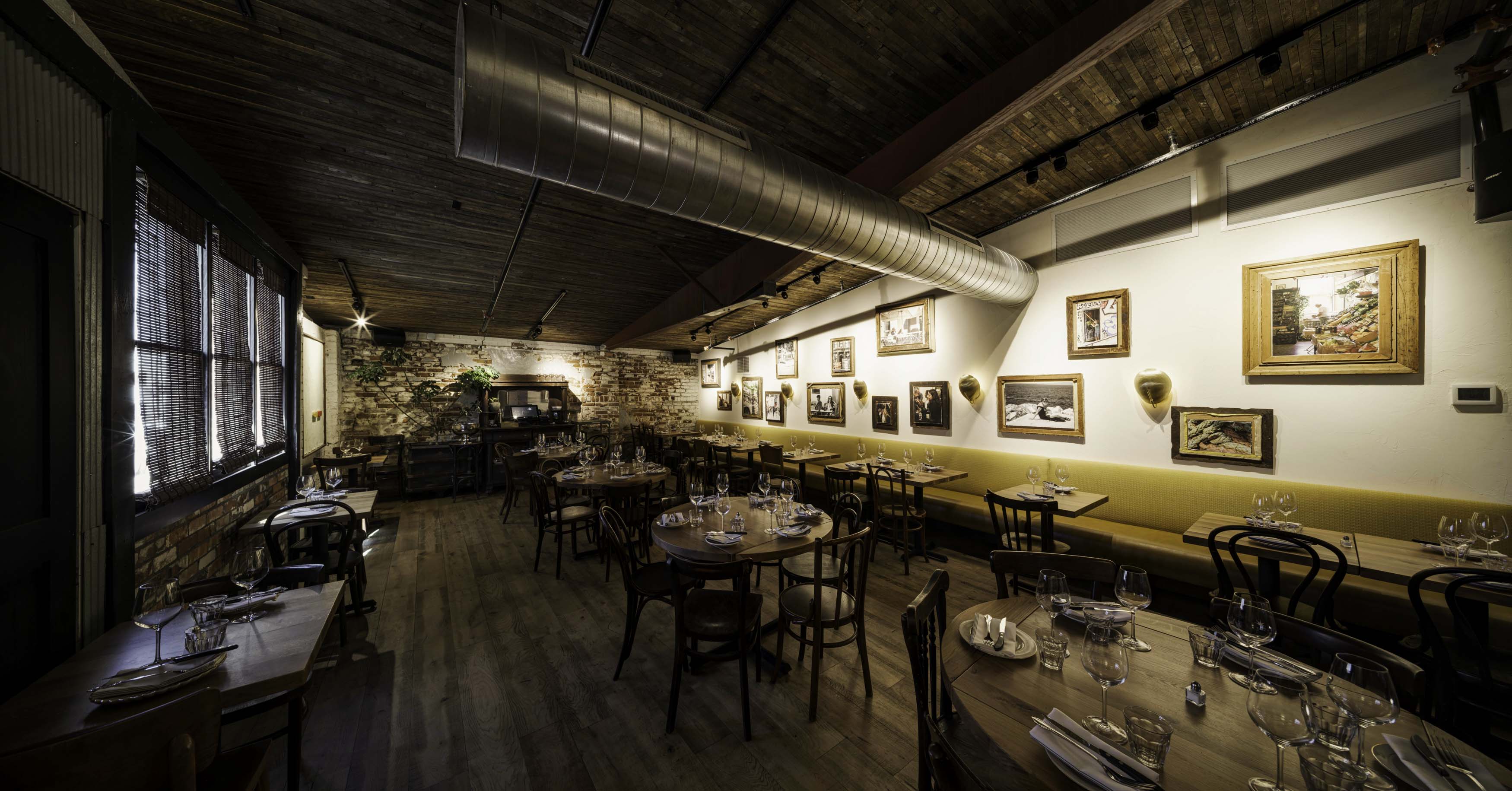 Cozy rustic restaurant interior with wooden floors, round and rectangular dining tables set with glassware, a yellow banquette along the wall, framed artworks, and exposed ductwork on a wooden ceiling.