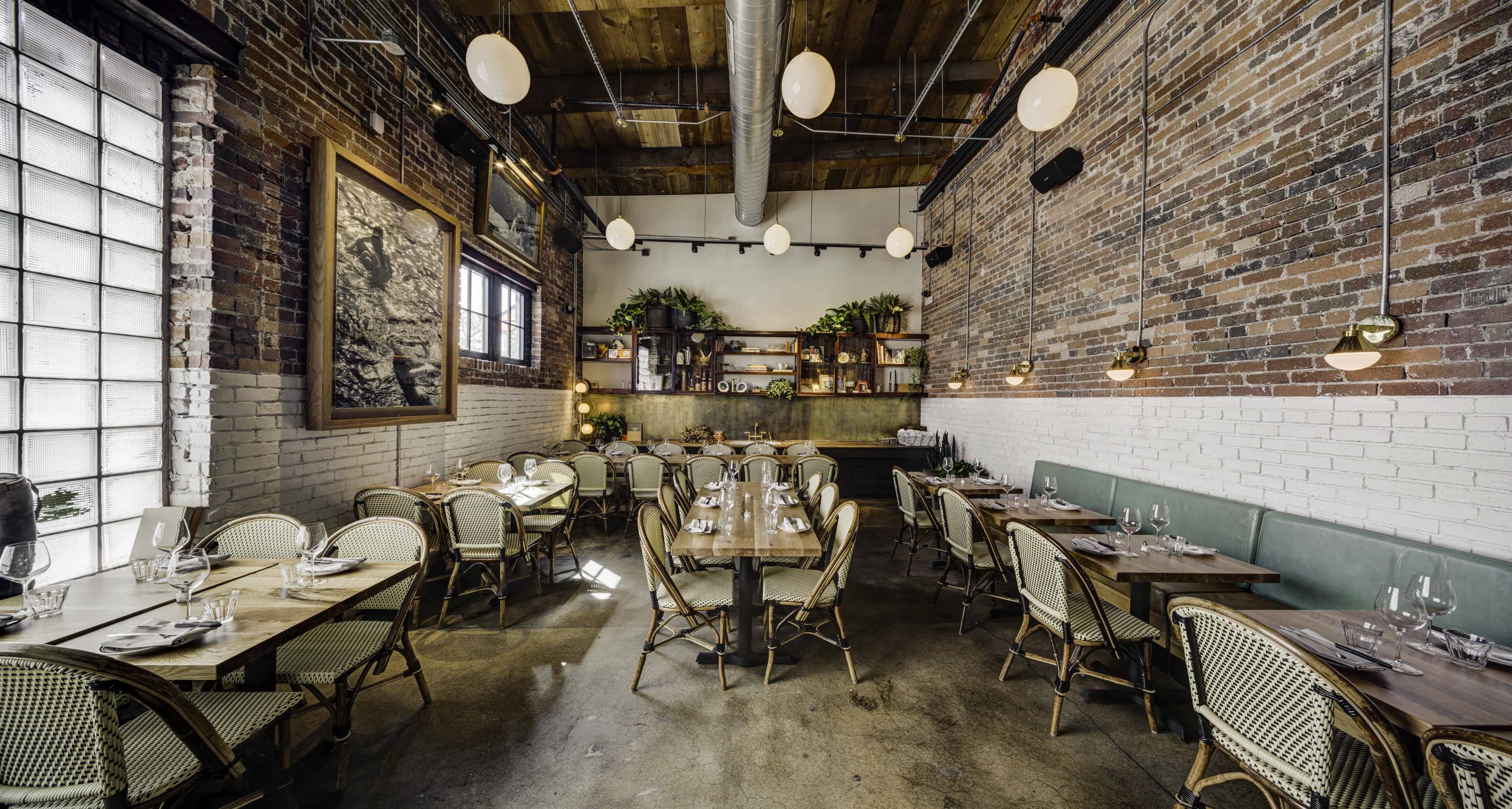 Modern rustic restaurant interior with exposed brick walls, wooden ceiling, hanging globe lights, wicker chairs, and neatly set wooden tables.