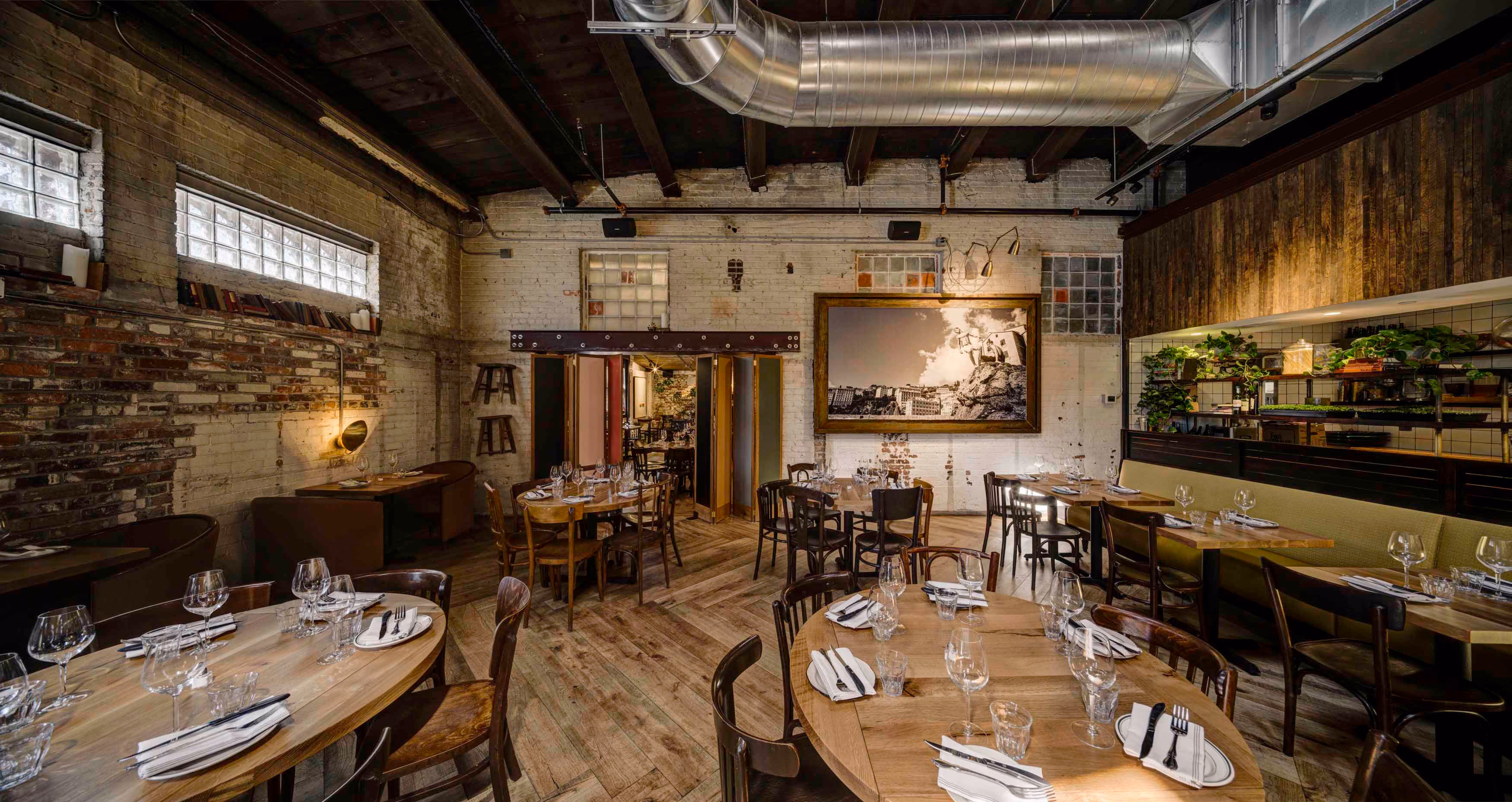 Rustic restaurant interior with wooden floors, round tables set with glassware, exposed brick walls, and greenery on shelves.