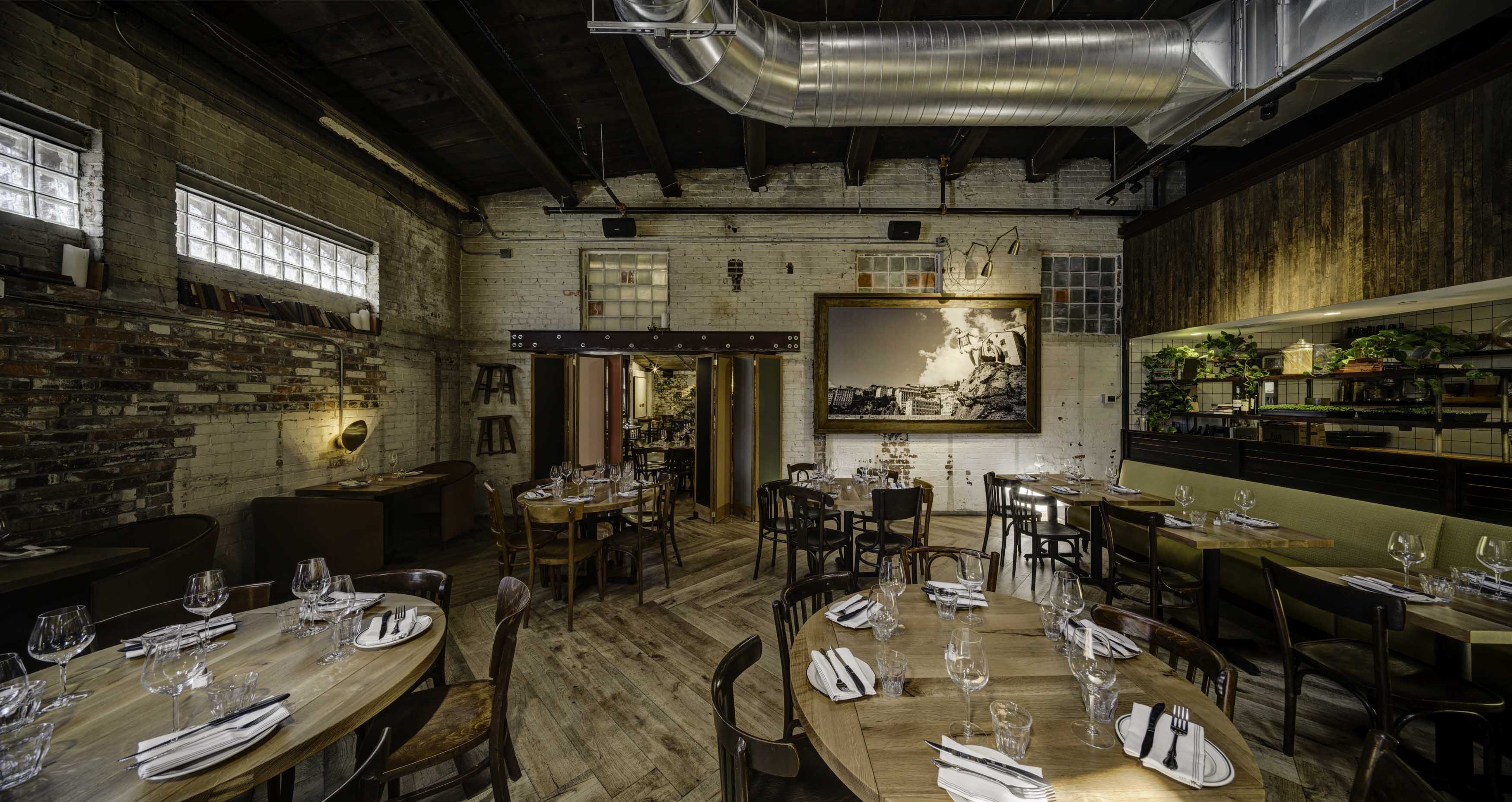 Rustic restaurant interior with wooden floors, round tables set with glassware, exposed brick walls, and greenery on shelves.