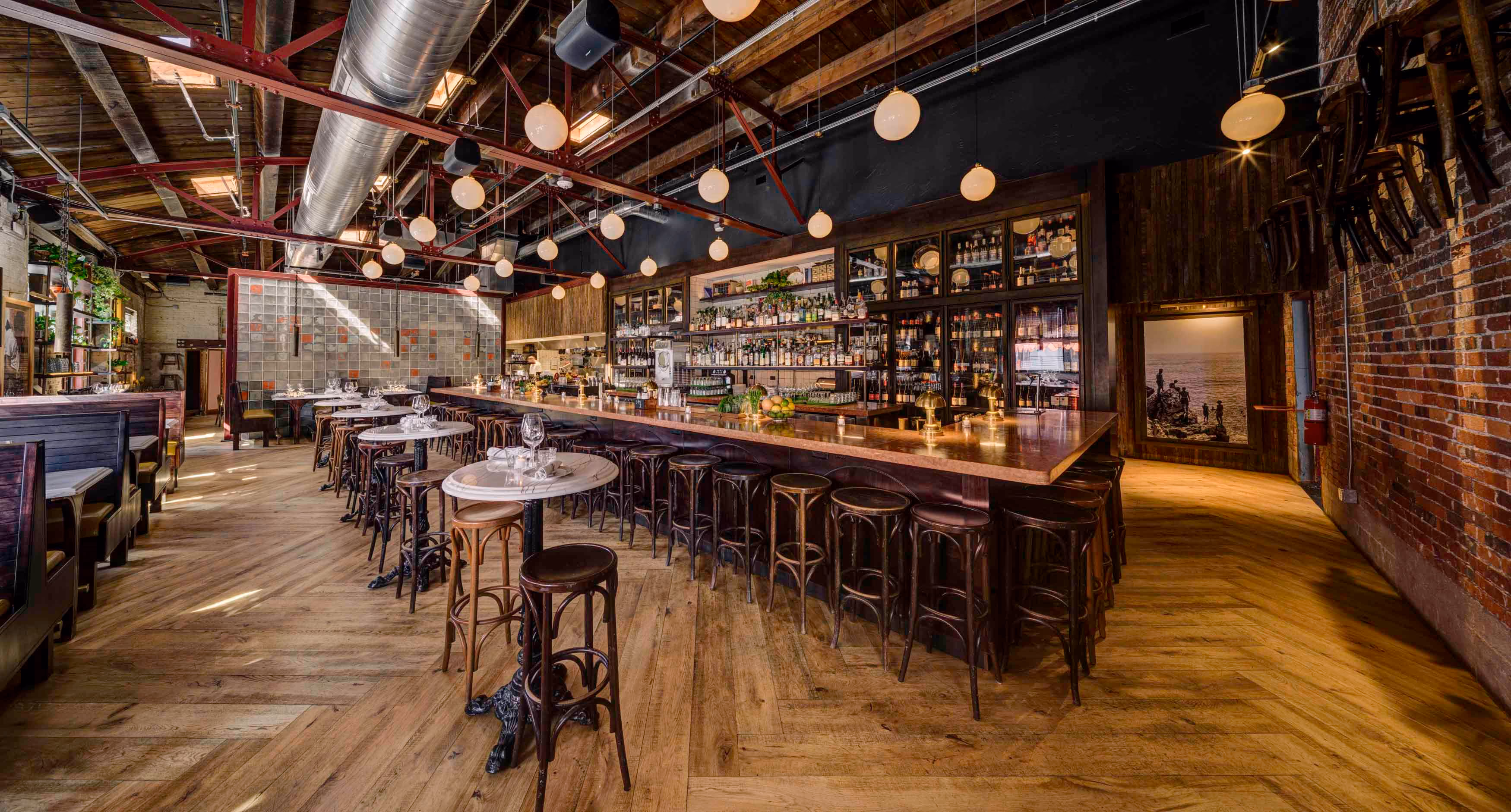 Spacious rustic bar and restaurant interior with wooden floors, a long bar counter with stools, marble tables, hanging globe lights, and exposed ceiling beams.