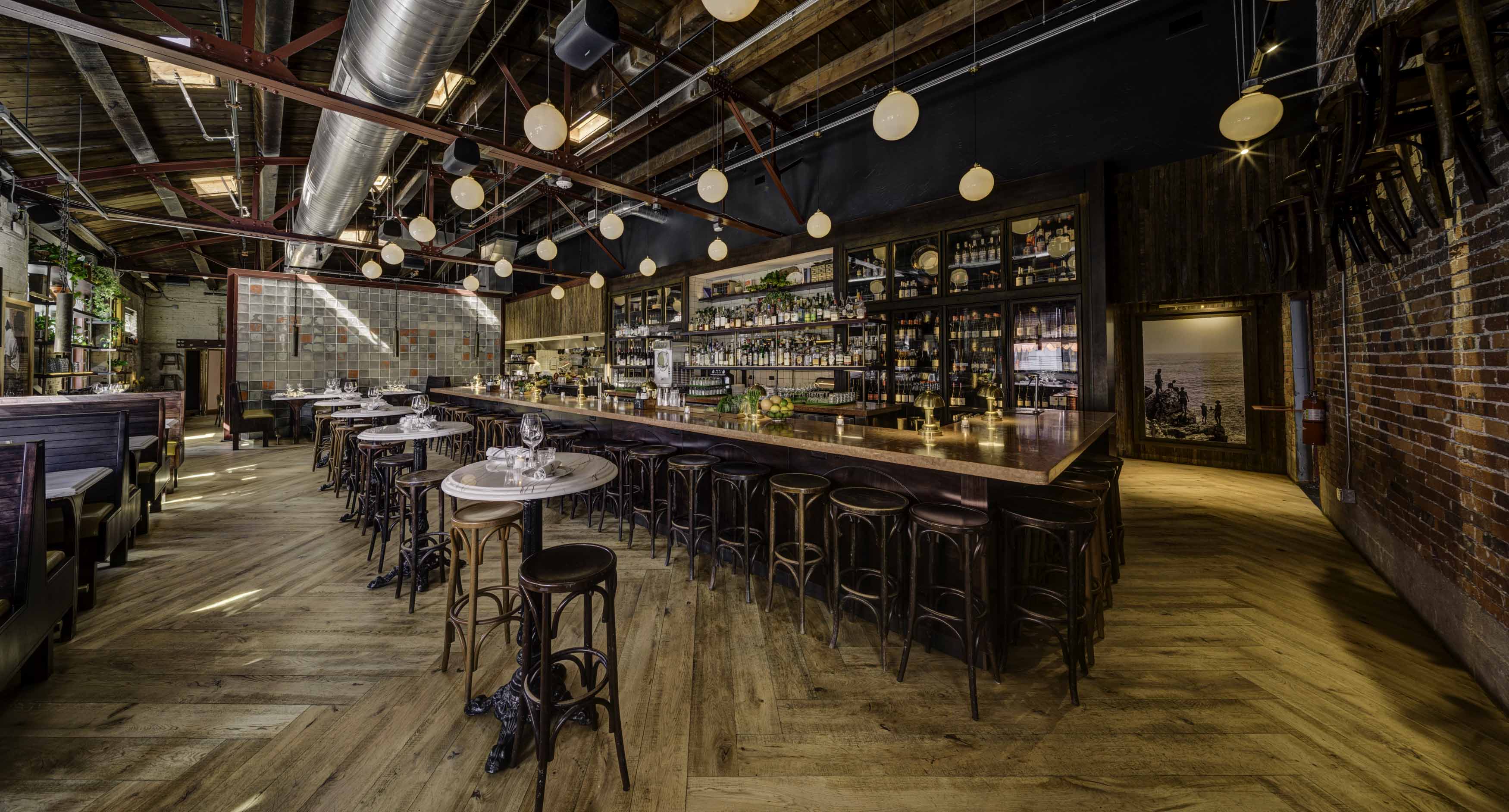 Spacious rustic bar and restaurant interior with wooden floors, a long bar counter with stools, marble tables, hanging globe lights, and exposed ceiling beams.