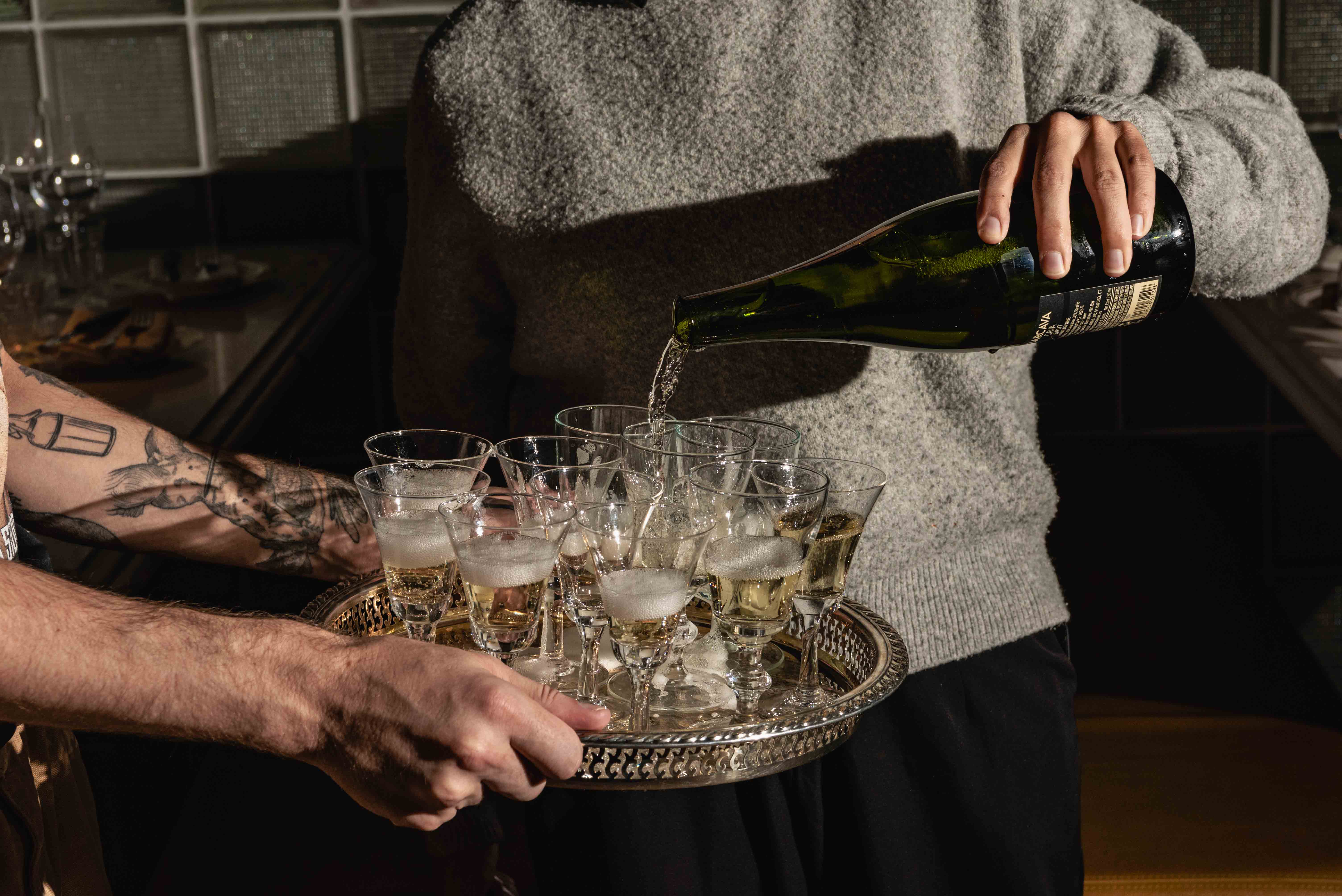 Person pouring sparkling wine into multiple glasses on a silver tray held by another person with a tattooed arm.
