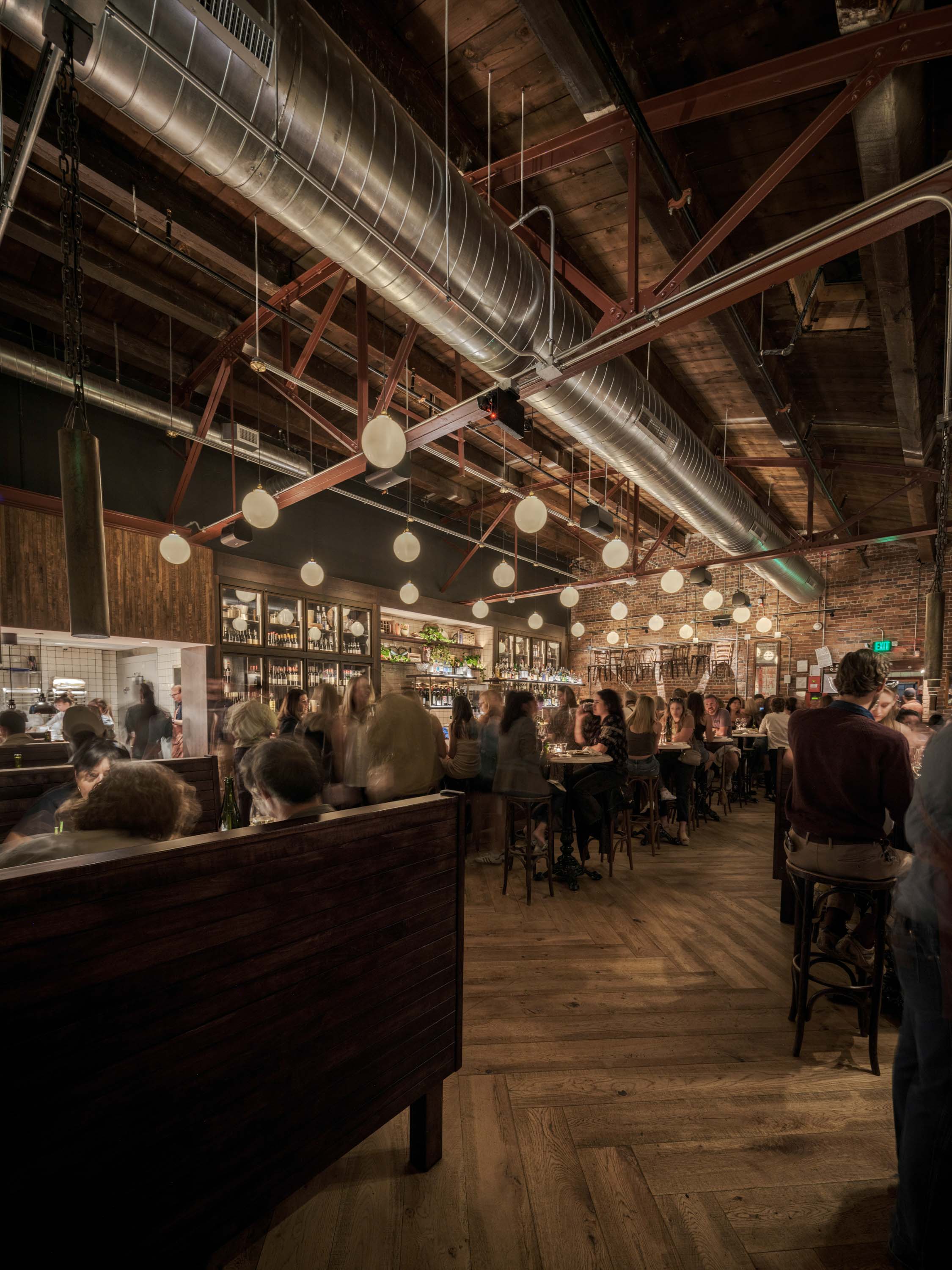 Interior of a crowded bar with wooden floors, exposed brick walls, hanging globe lights, and visible metal ductwork on a high ceiling.