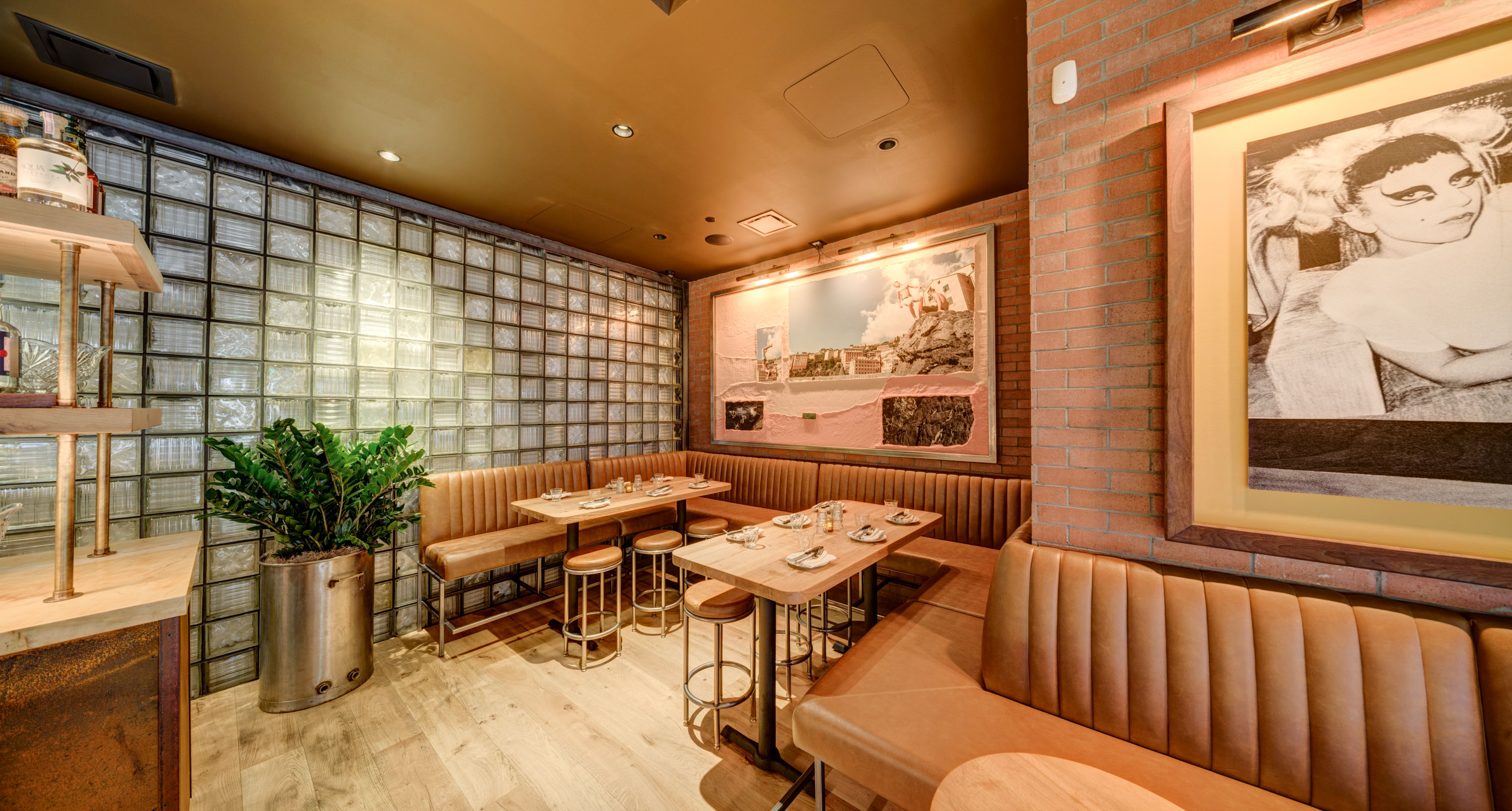 Cozy restaurant corner with tan leather booths, round stools, wooden tables set with glasses and napkins, and framed artwork on brick and glass block walls.