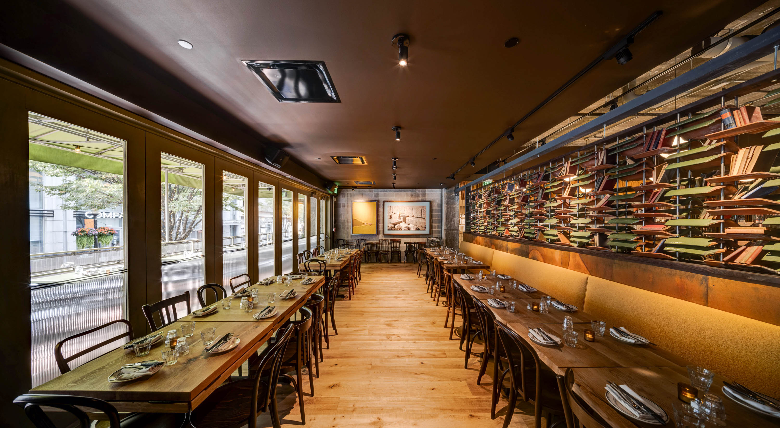 Interior of a modern dining area with long wooden tables set with plates and glasses, cushioned bench seating on one side, and decorative shelving with books along the wall.