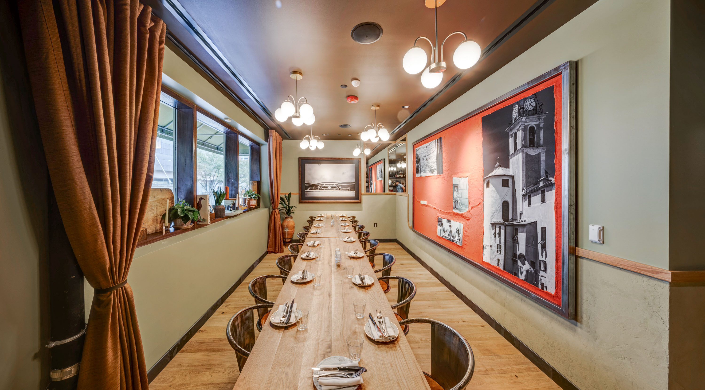 Long wooden dining table set with plates, napkins, and cutlery in a cozy room with large windows, warm lighting, and wall art featuring black and white photos framed by red background.