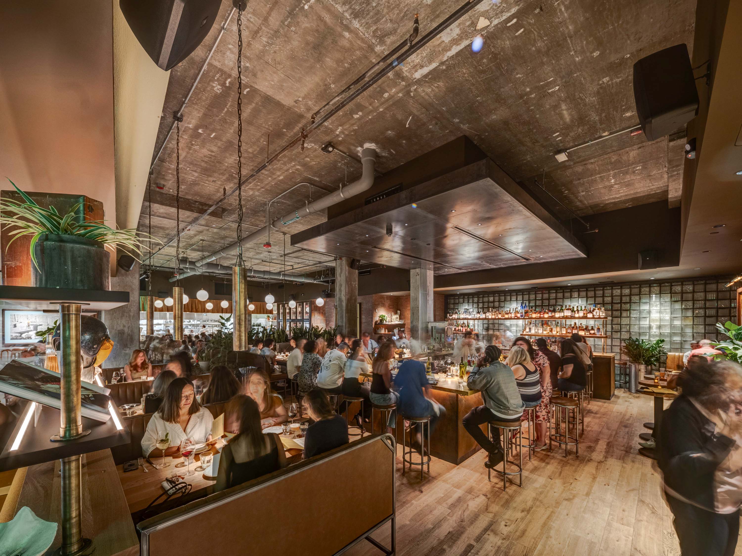 Busy modern bar and restaurant interior with patrons dining and socializing under industrial-style ceiling and warm lighting.