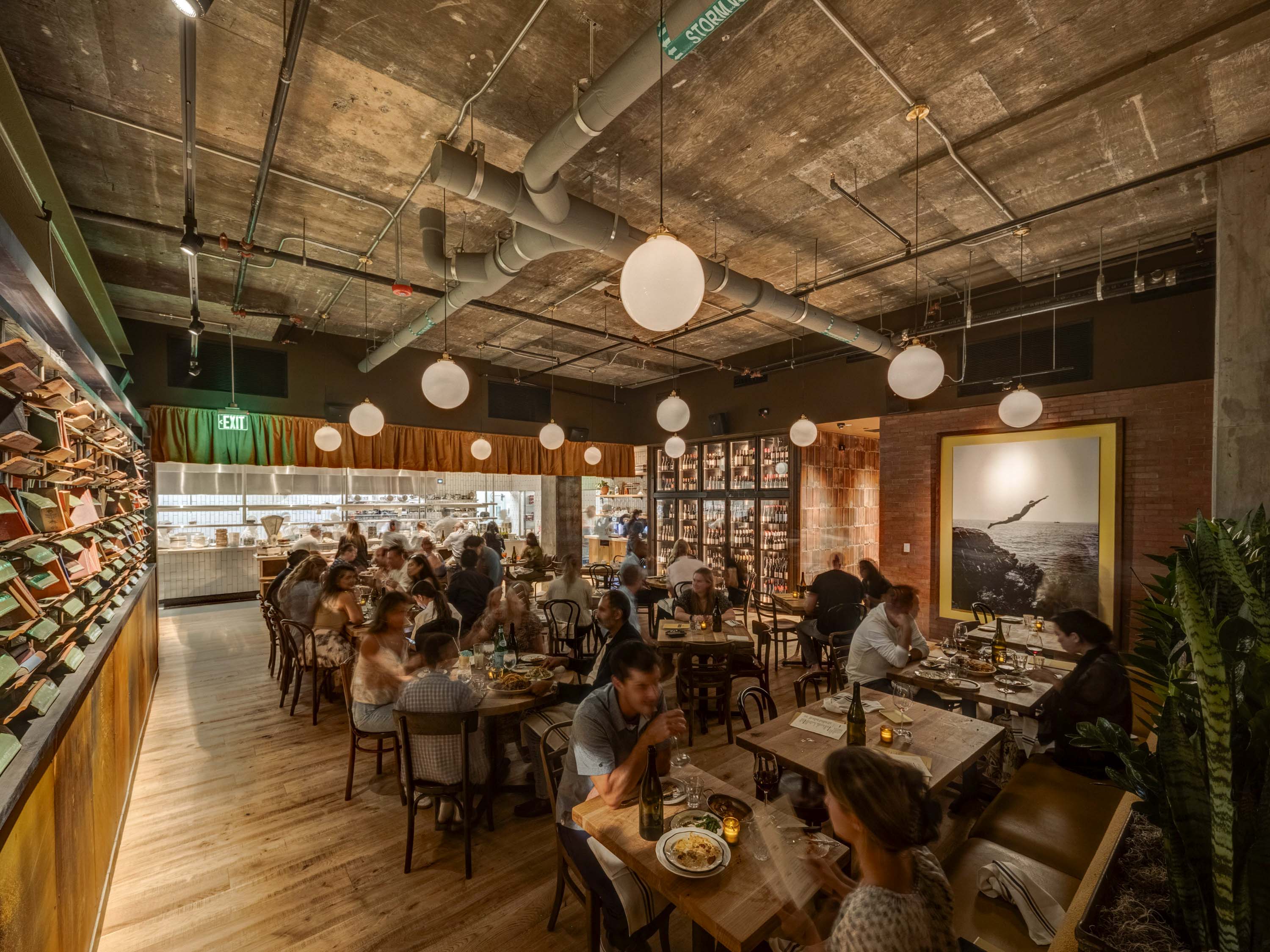 Cozy restaurant interior with exposed concrete ceiling, round pendant lights, wooden tables, and diners enjoying meals.