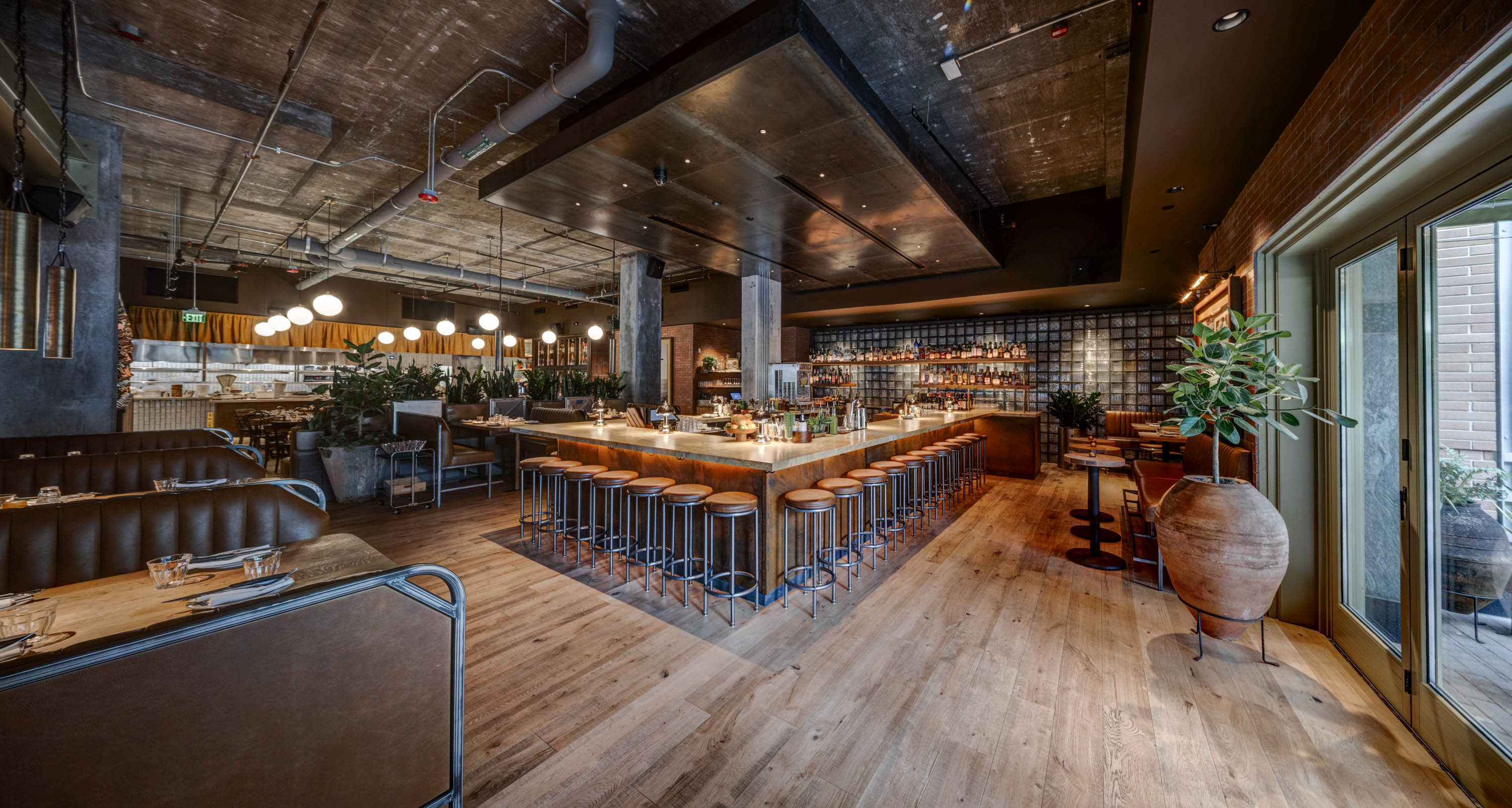 Modern restaurant interior with a central U-shaped bar surrounded by high stools, seating booths, wooden floors, and large potted plants near glass doors.