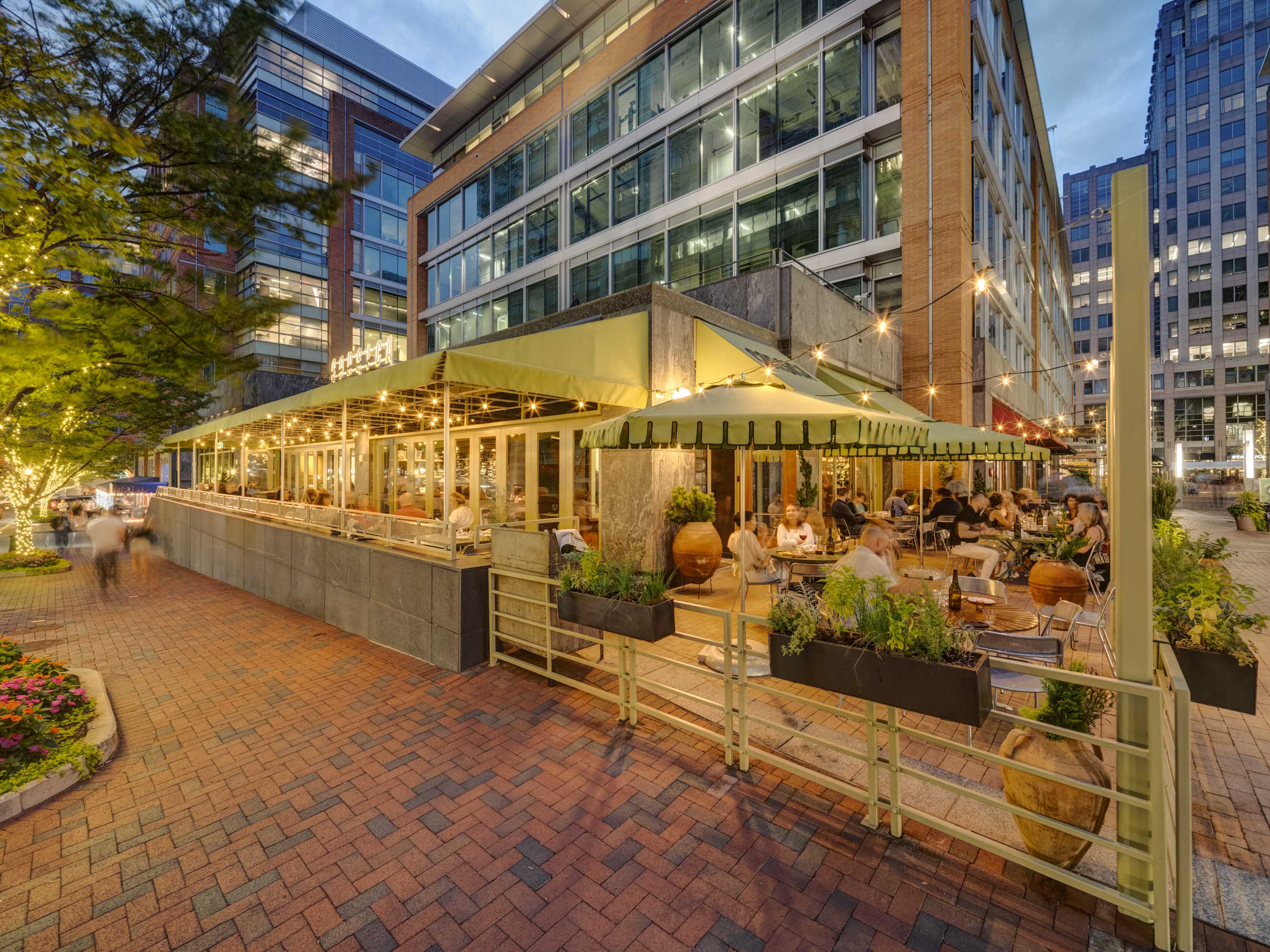Outdoor restaurant patio with patrons dining under green umbrellas and string lights in an urban setting at dusk.