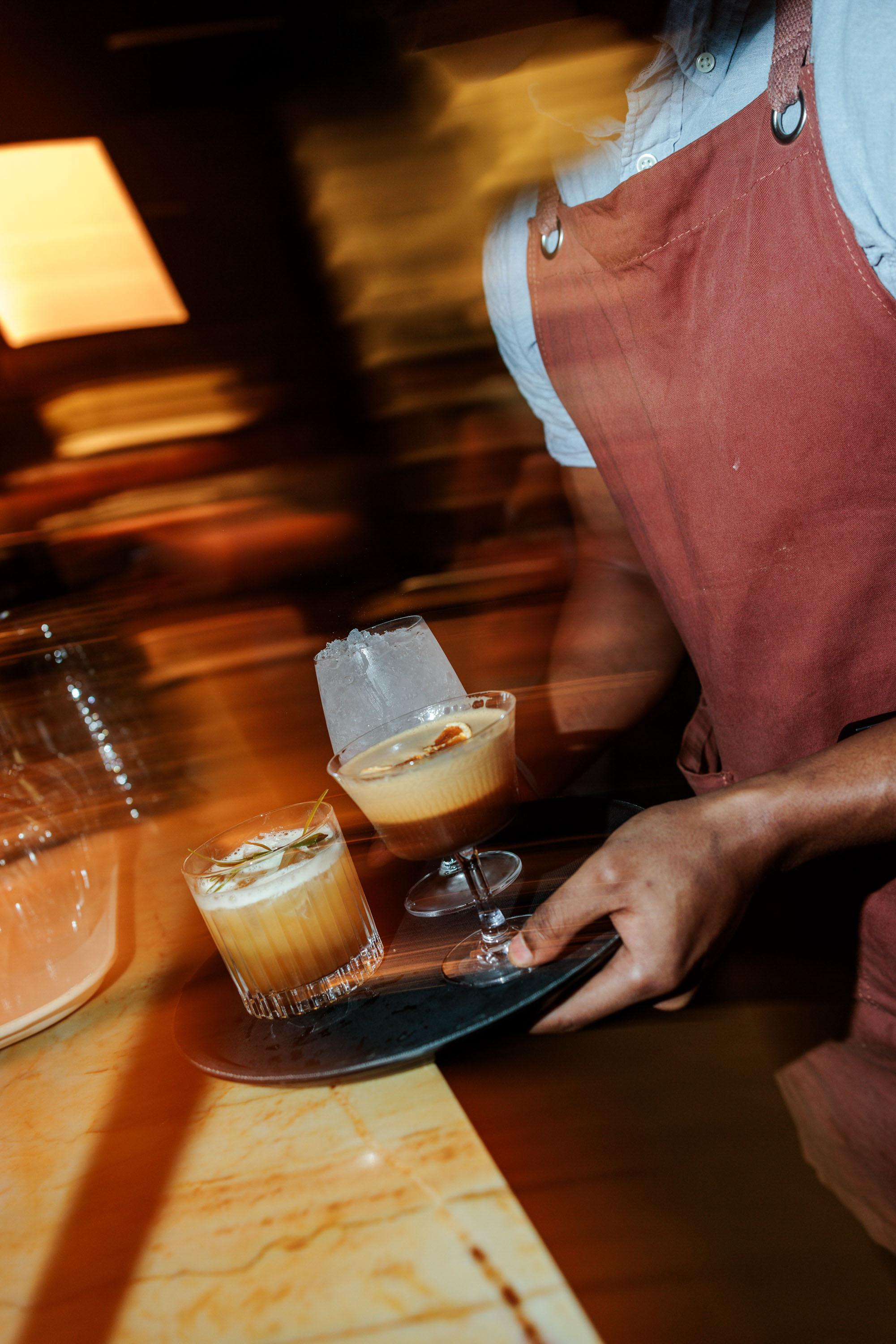 Bartender wearing a red apron holding a tray with three cocktails, including a glass of ice and two garnished drinks, near a bar counter.
