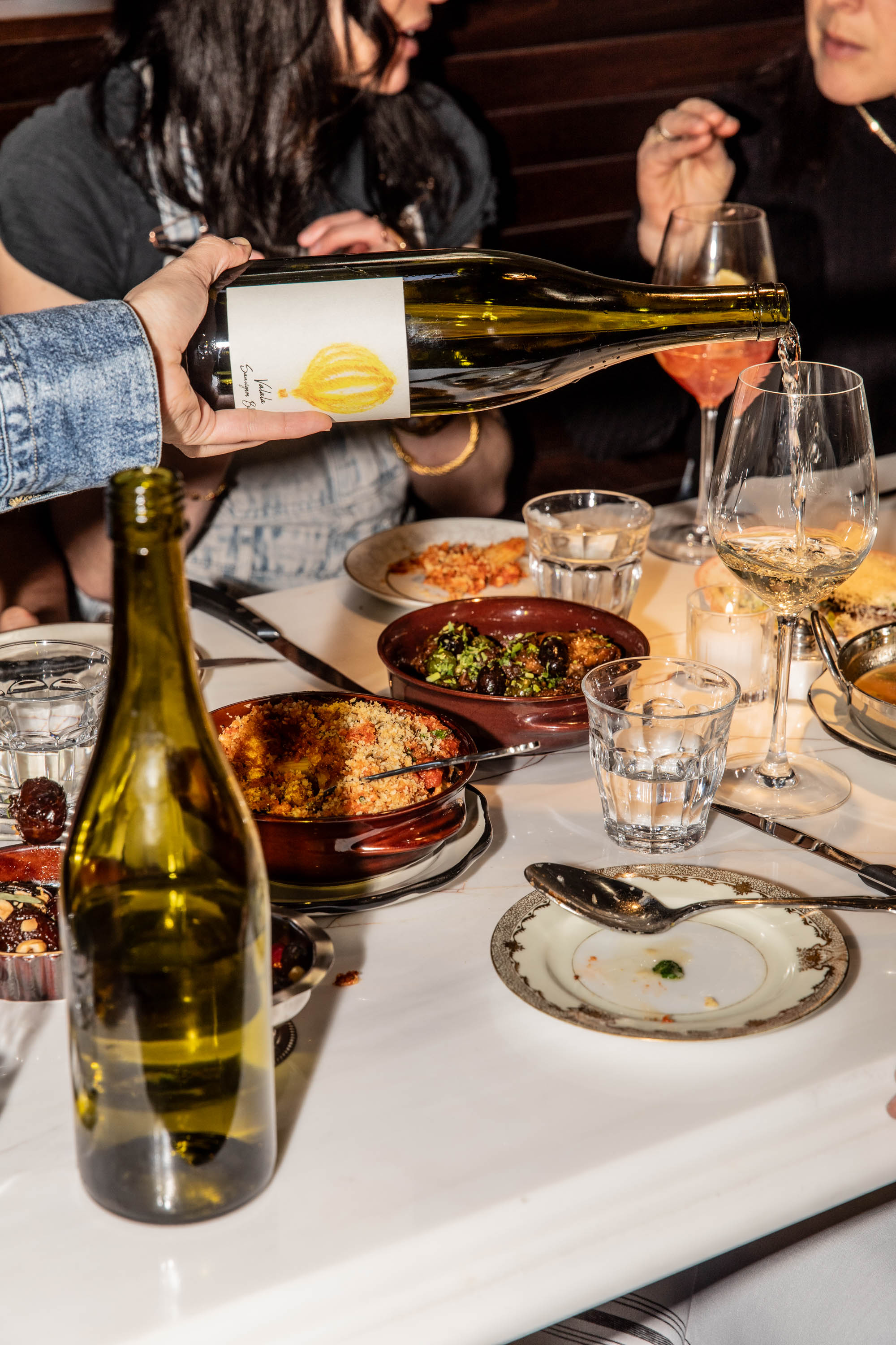 Hand pouring white wine from a bottle into a glass at a dinner table with plates of food and people in the background.
