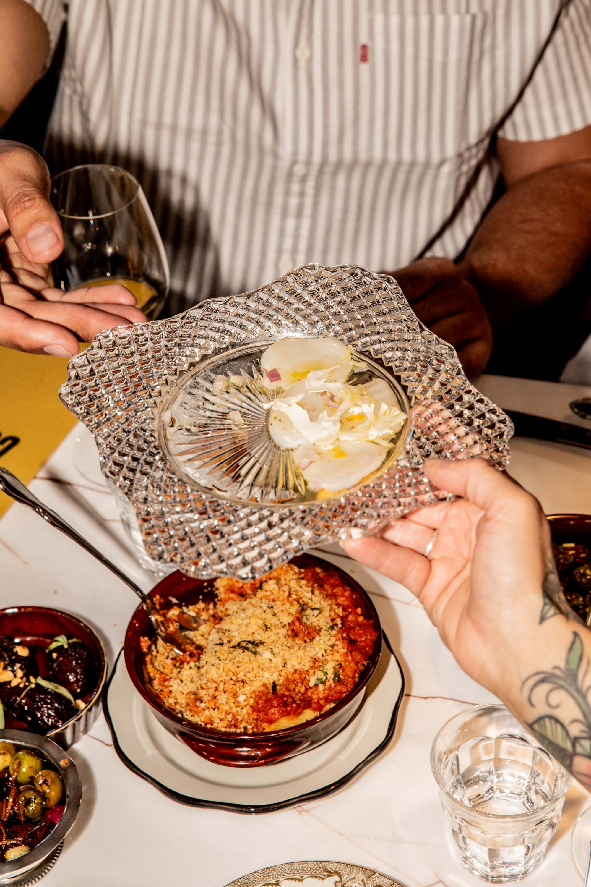 Two hands lifting a decorative glass lid off a red dish filled with breadcrumb-topped baked food on a white table with small bowls of olives and other dishes nearby.