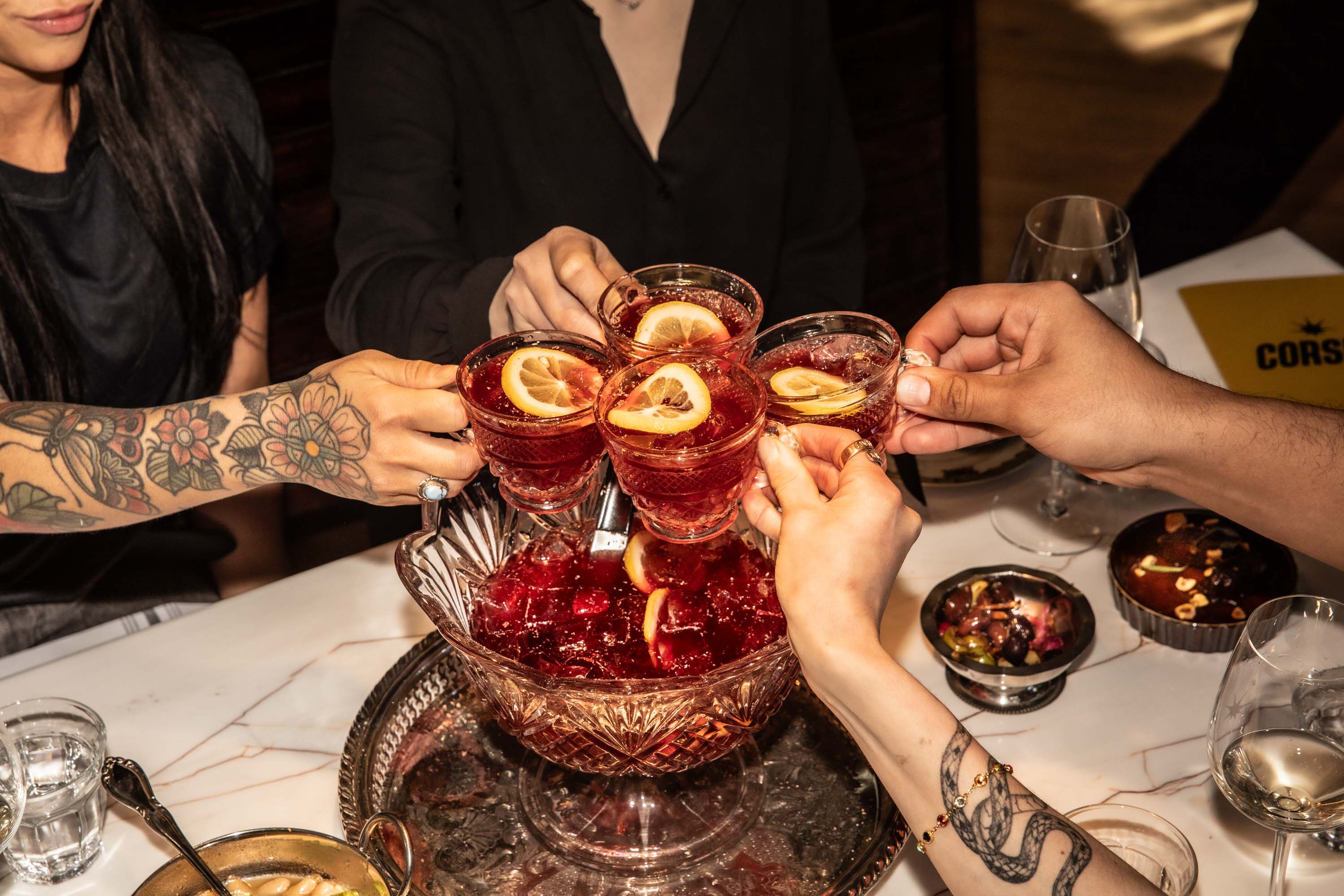 Four hands clinking crystal glasses filled with red cocktail garnished with lemon slices over a punch bowl on a marble table.