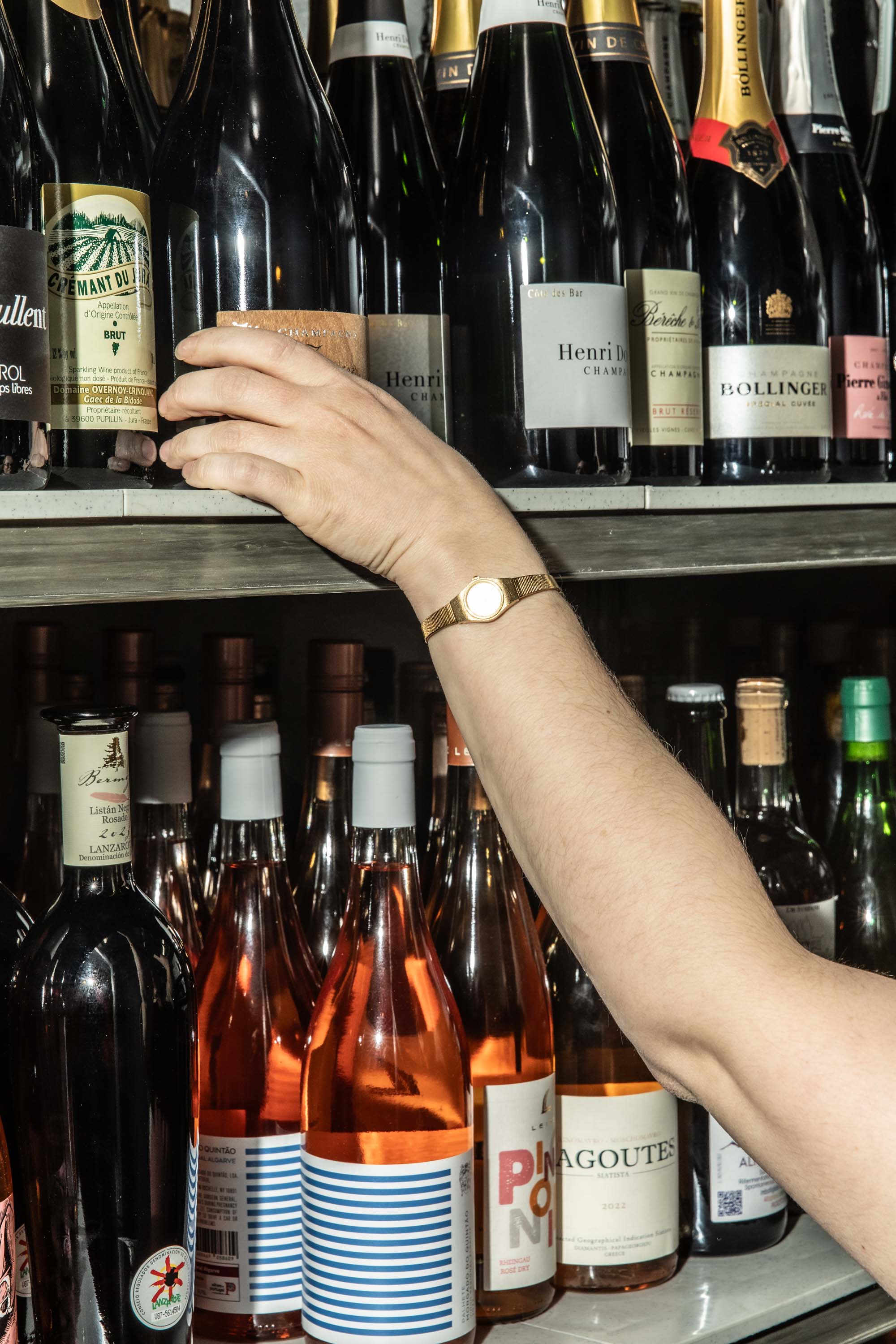 Person wearing a gold watch reaching for a bottle of sparkling wine on a shelf filled with assorted wine and champagne bottles.