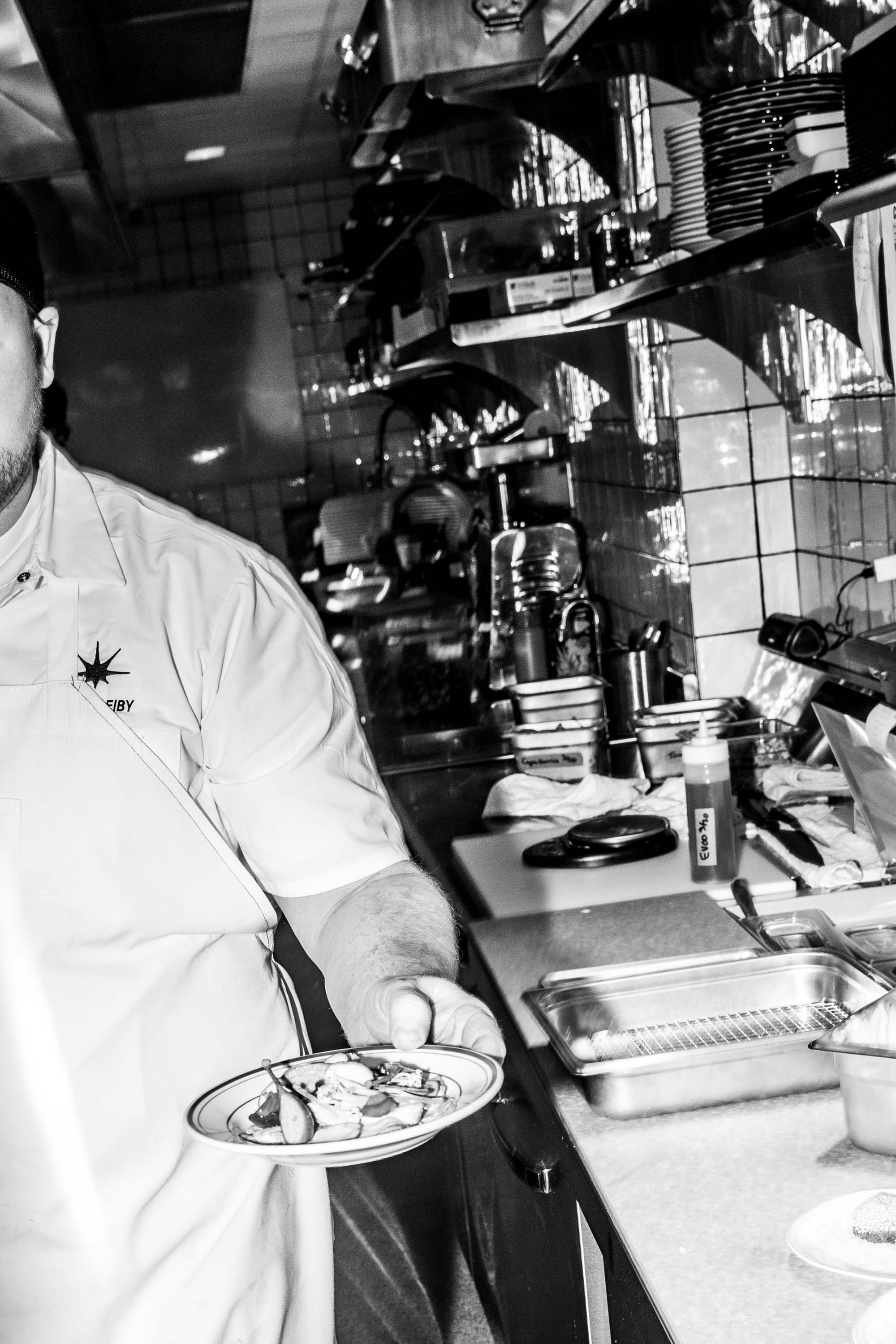 Chef in a kitchen holding a plate of food with cooking utensils and shelves in the background.