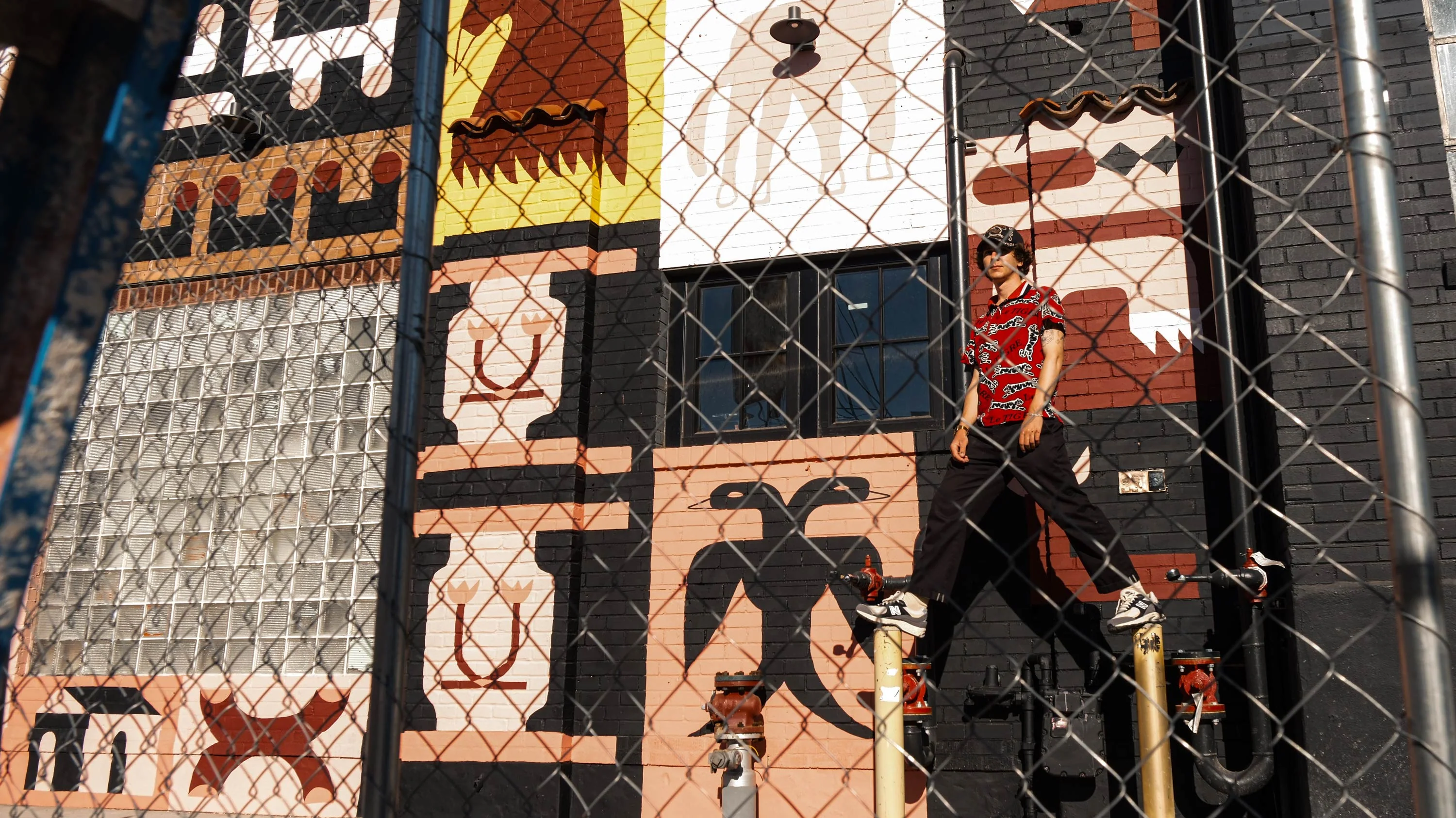 Person in red patterned shirt and black pants standing on utility pipes in front of a brick wall with colorful abstract mural.