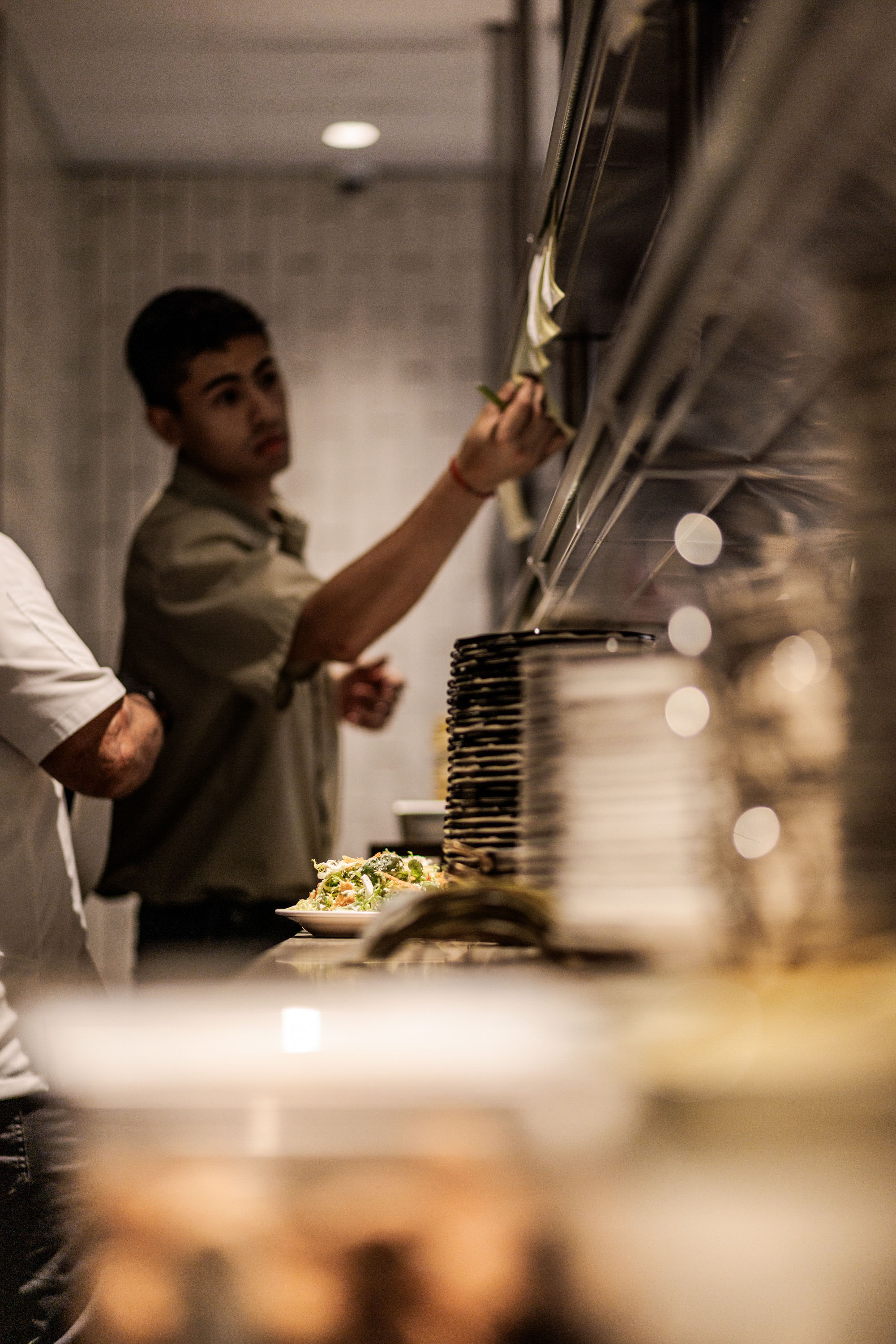 Young man reaching for a note on a kitchen order wall with a stack of plates and prepared food on a counter.