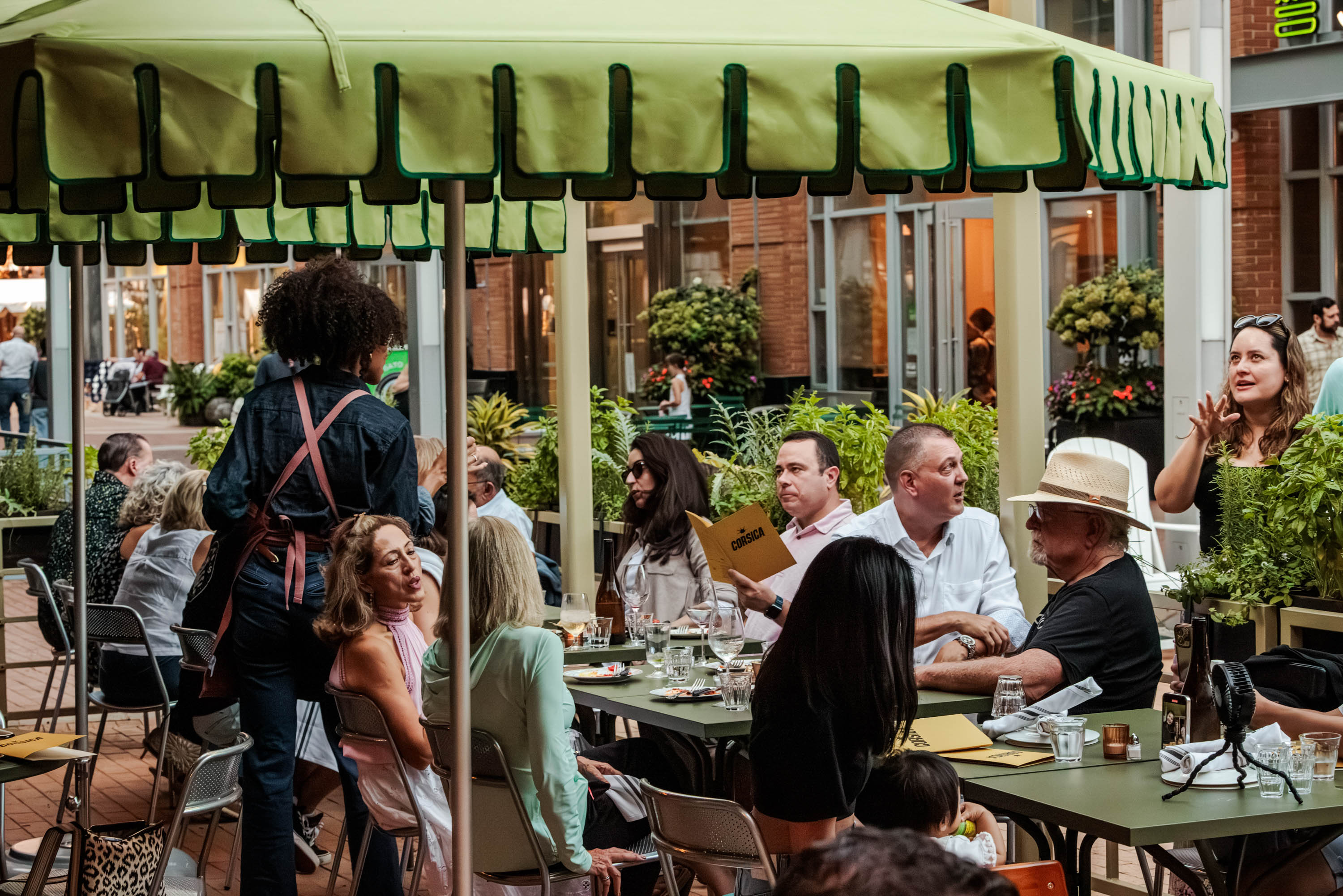 People dining and conversing under a large green patio umbrella at an outdoor restaurant with menus on the tables.