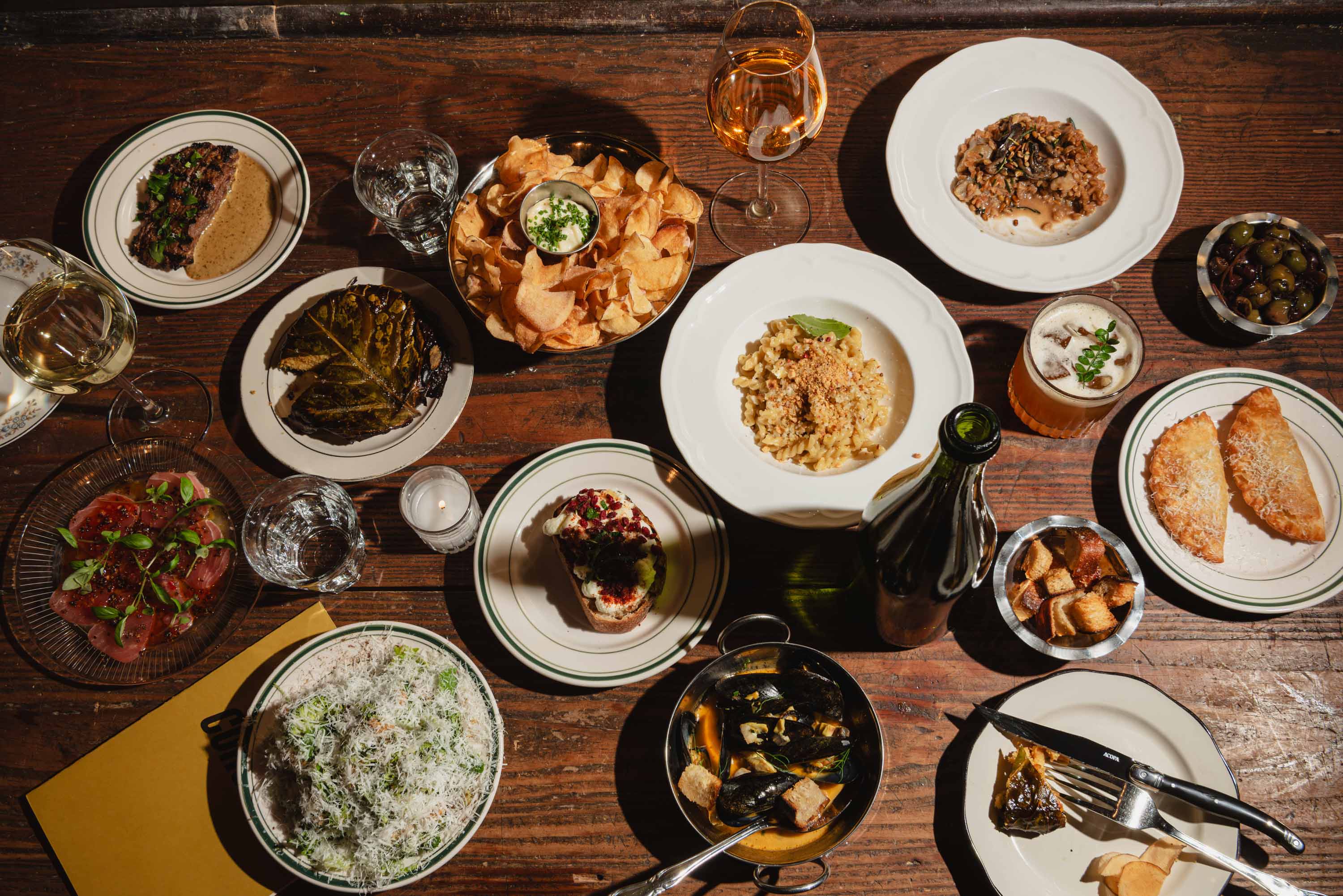 Table set with various dishes including pasta, mussels, salad with grated cheese, empanadas, olives, potato chips with dip, toasted bread with spread, sliced meat, and drinks including white and amber wine, and a cocktail.