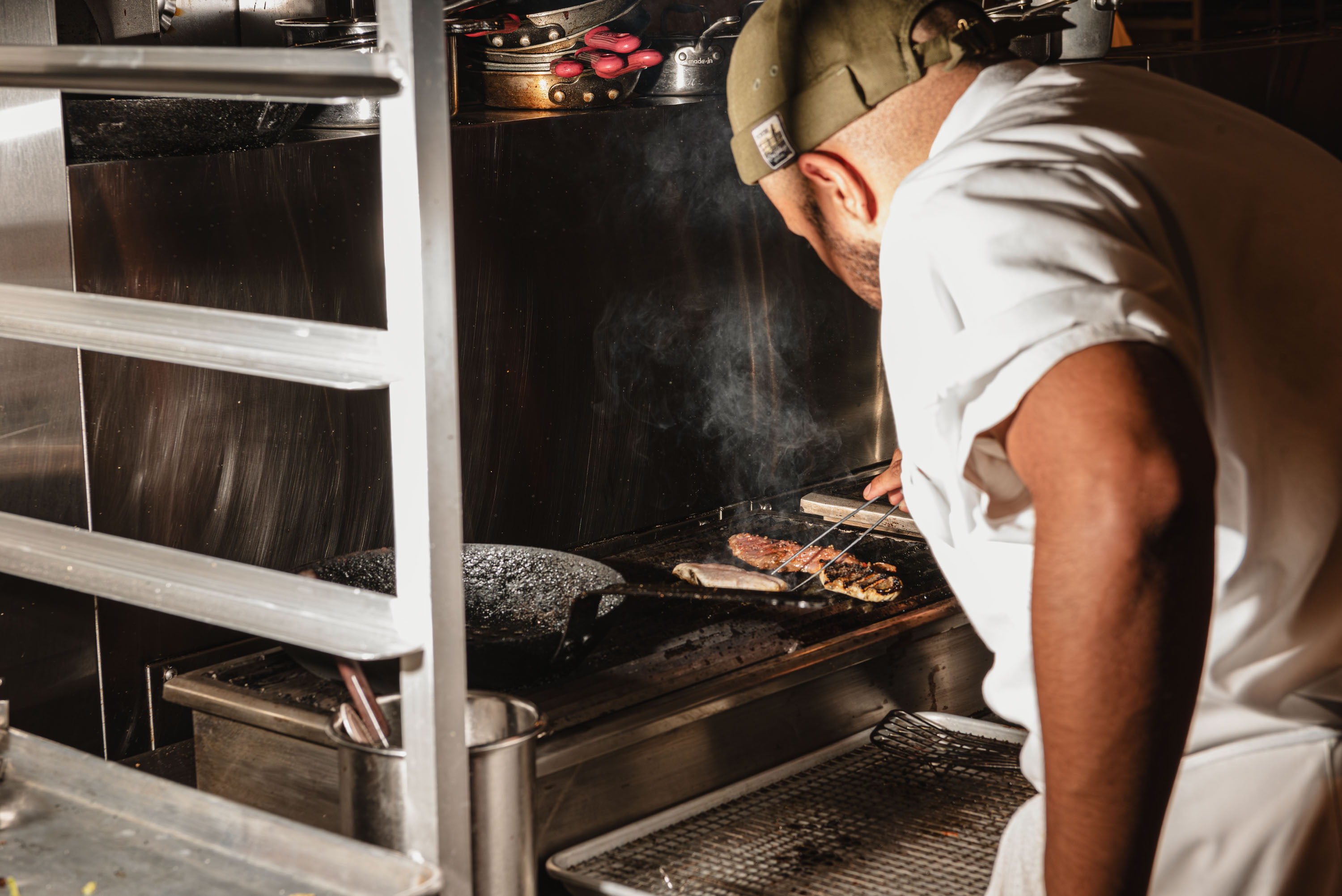 Chef wearing a cap grilling meat on a commercial stove in a kitchen.