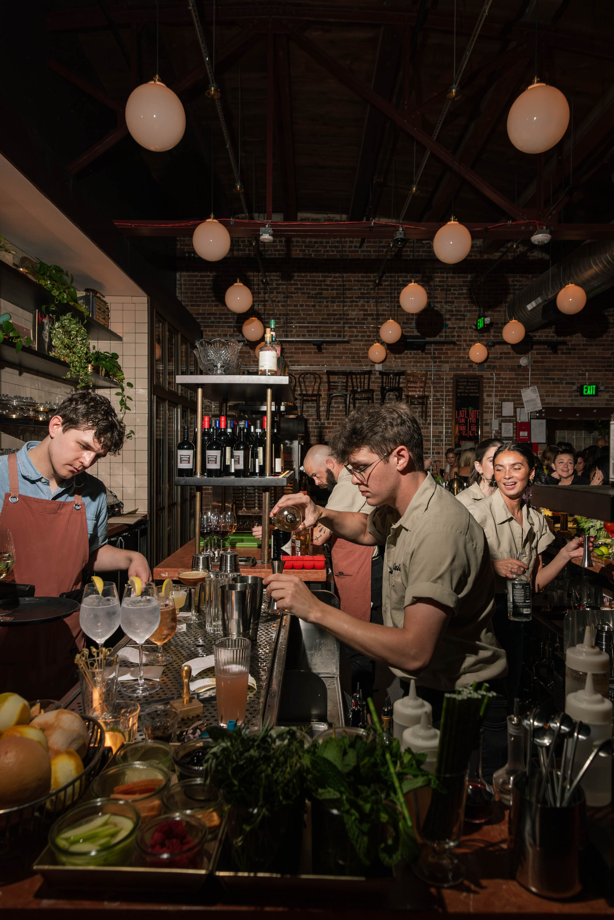 Bartenders preparing drinks behind a busy bar with hanging round lights and exposed brick walls.