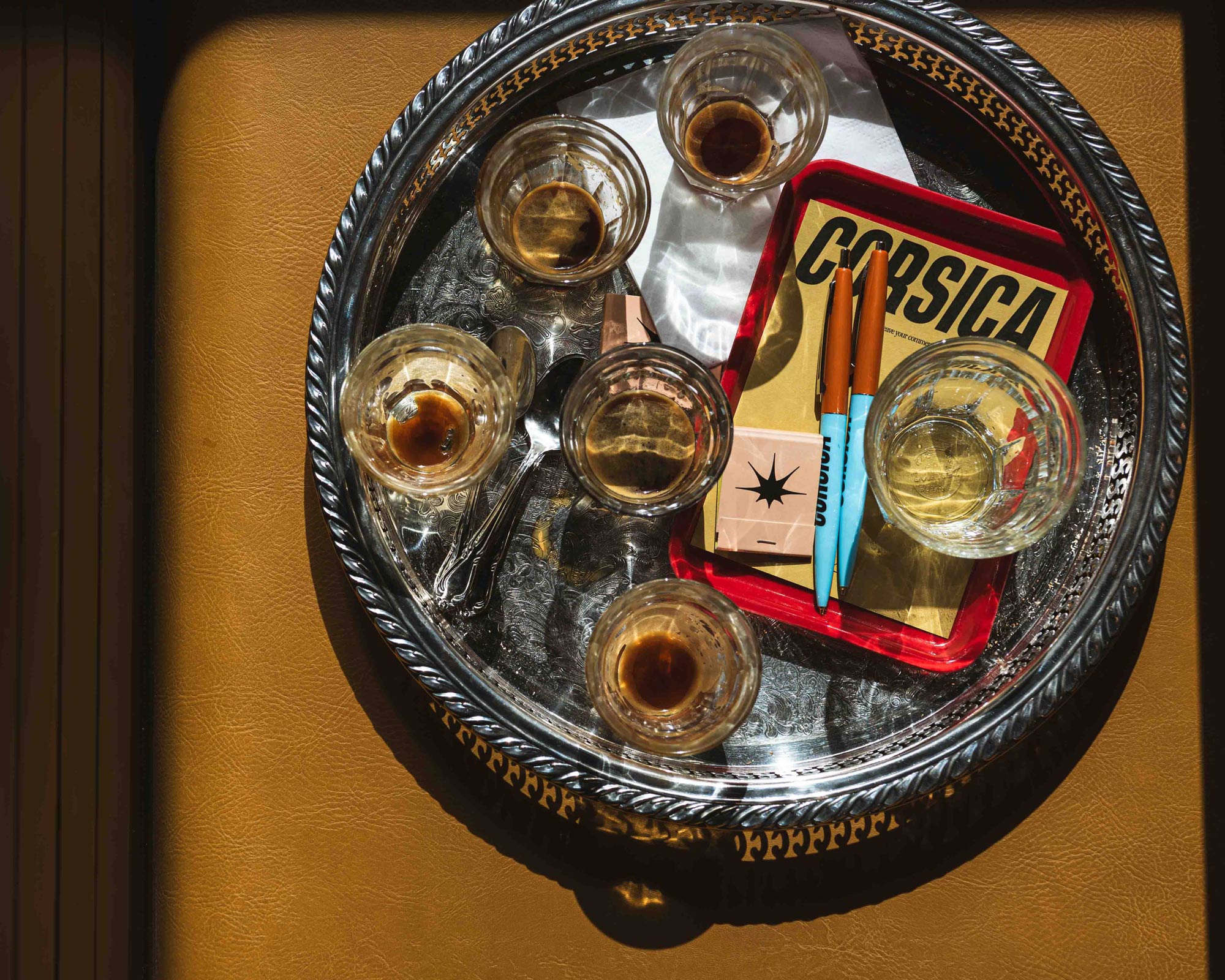 Silver tray on a brown surface holding five small glasses with espresso, two blue and orange pens, and a red-bordered book titled 'CORSICA'.