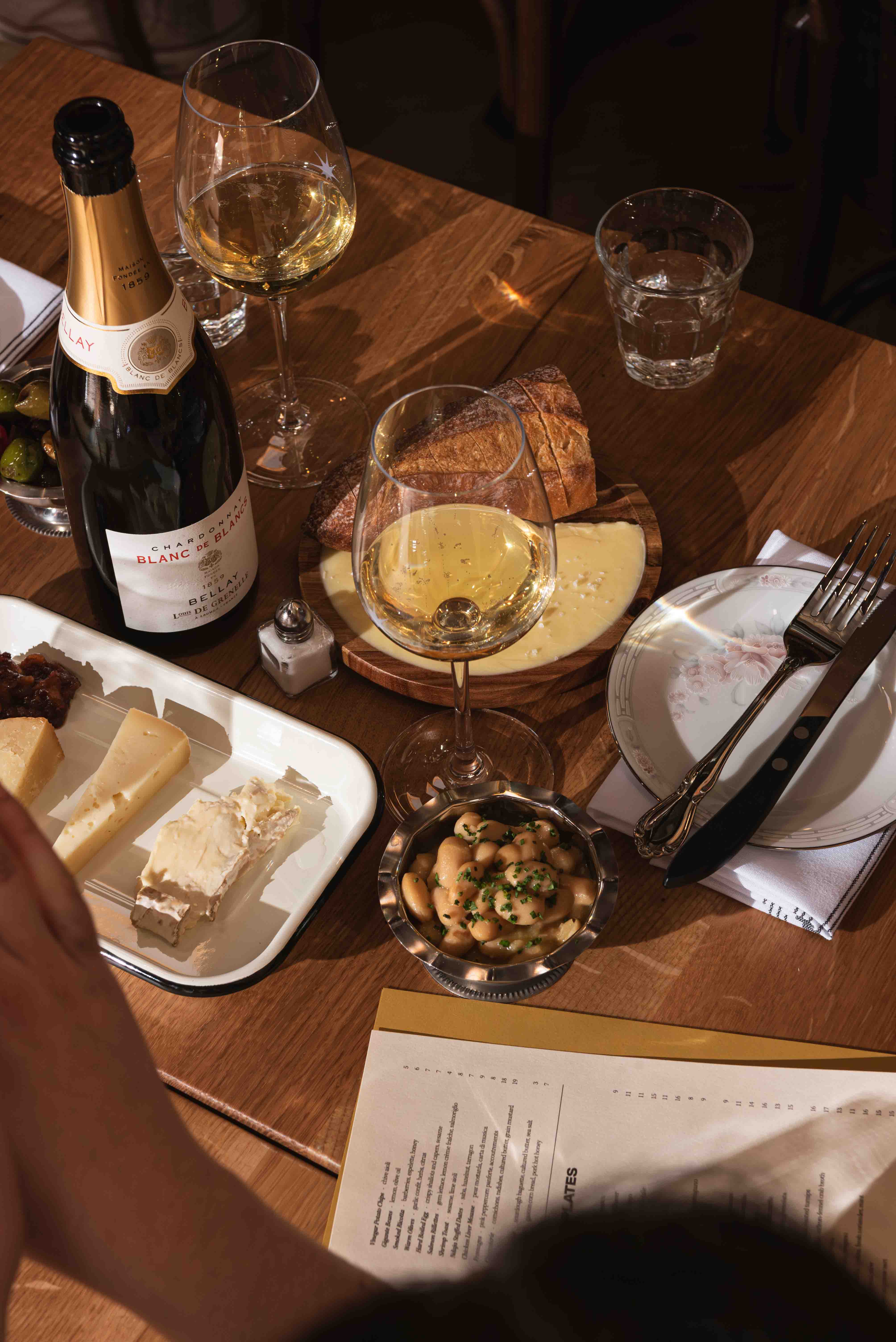 Table set with a bottle of Blanc de Blancs champagne, two glasses of white wine, a cheese platter with three kinds of cheese, bread, a bowl of white beans, a small dish of green olives, and a fork and knife on a plate.