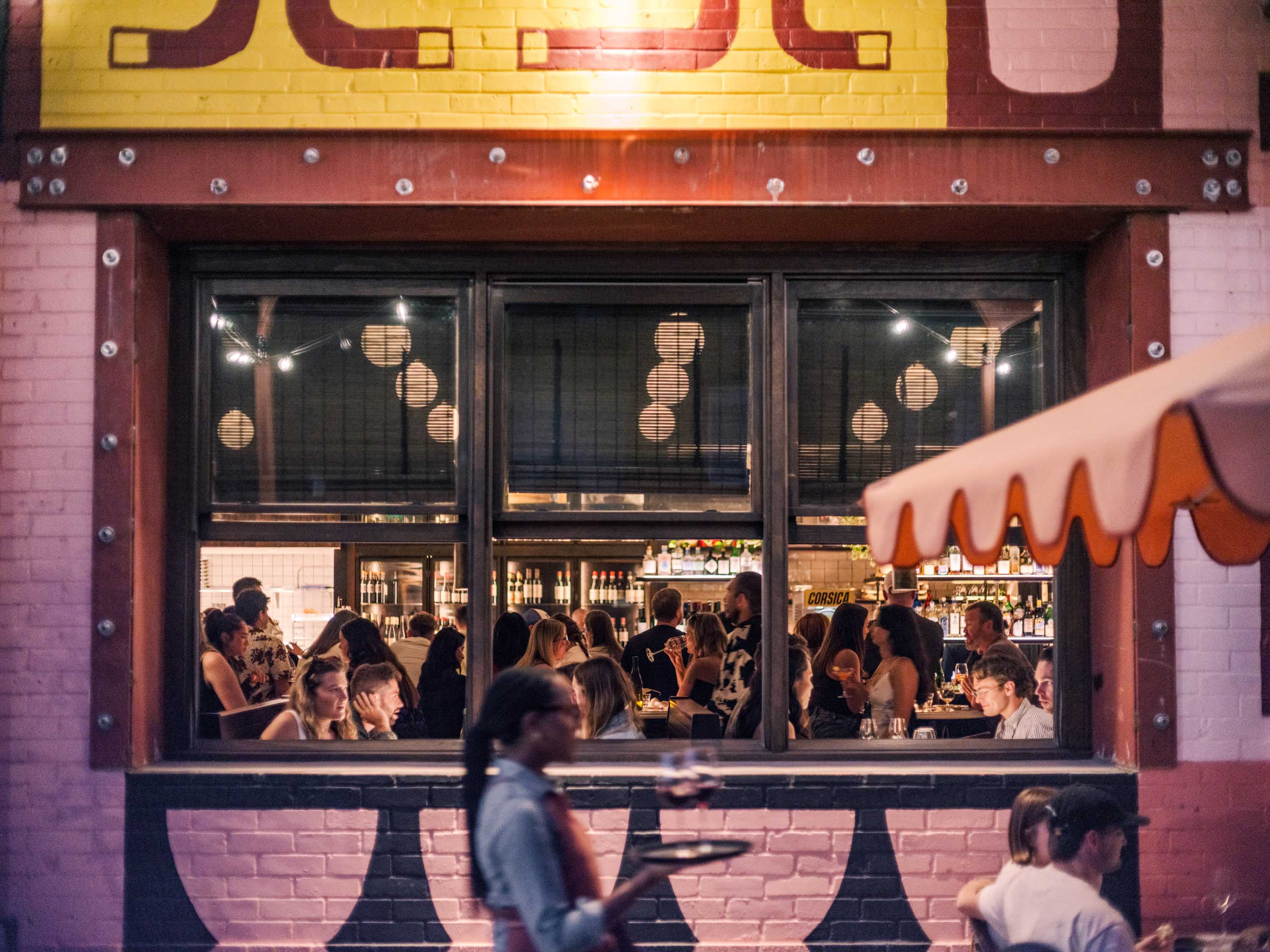 View through a large window of a crowded bar or restaurant with patrons socializing and a server carrying drinks outside.
