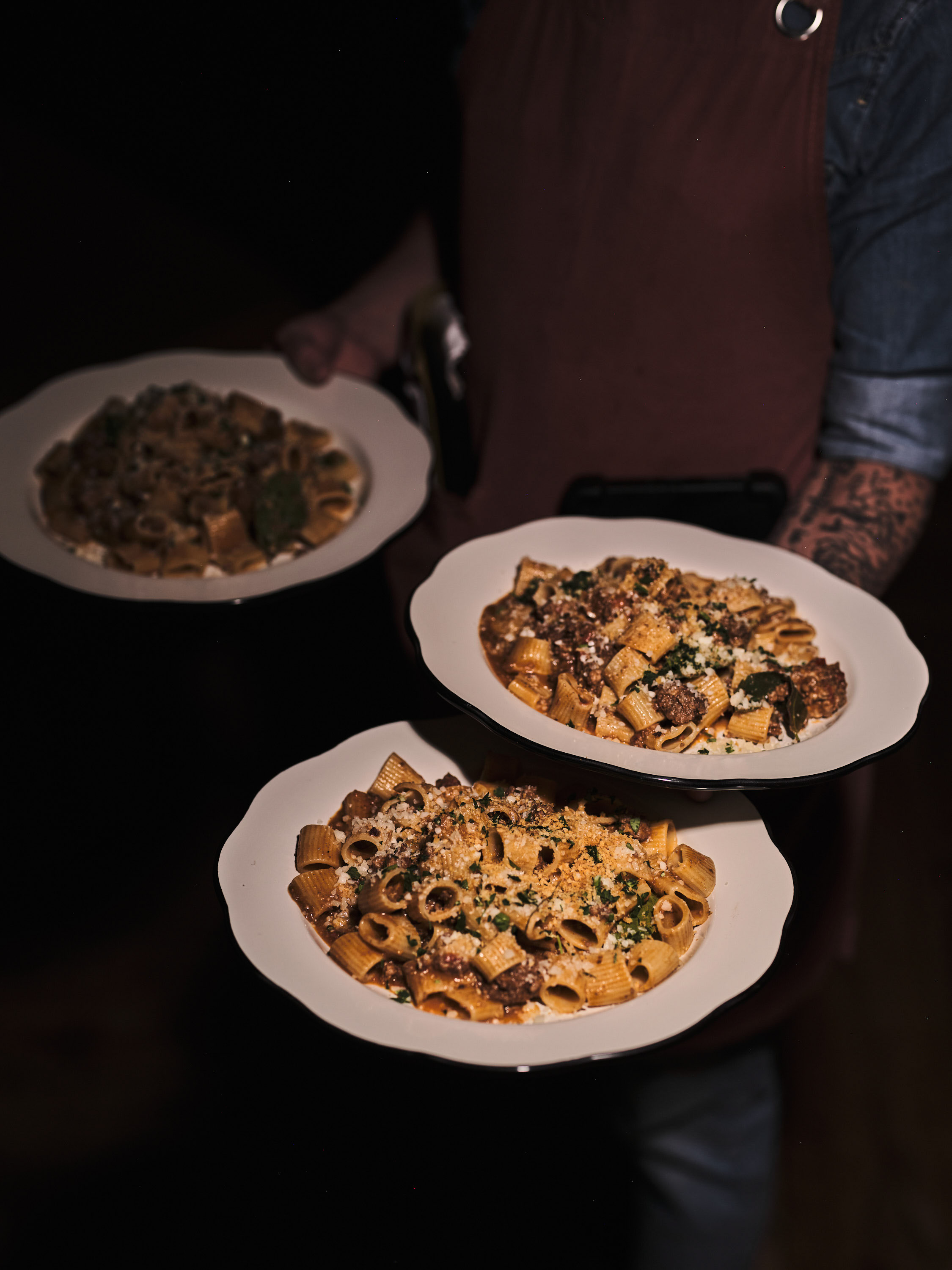 Person in a brown apron carrying three plates of pasta with sauce and grated cheese.