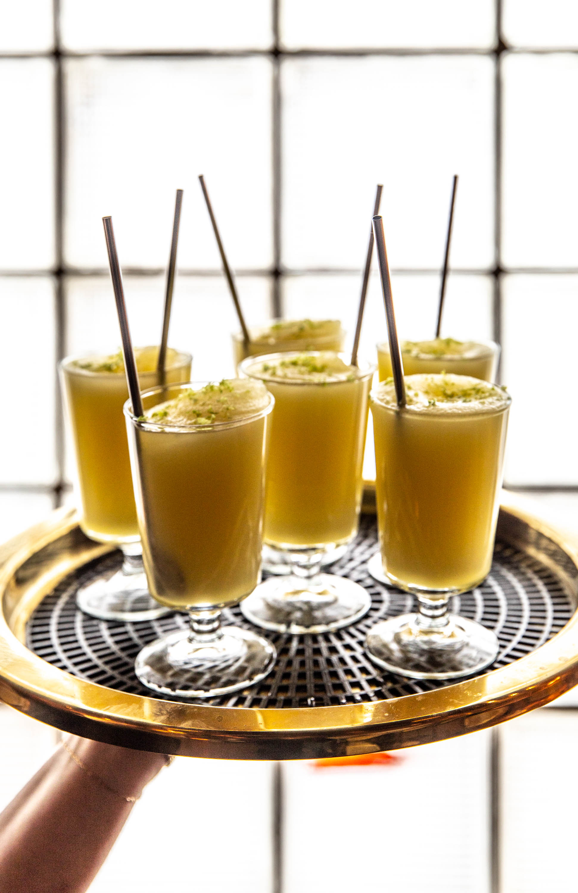 Tray held by a hand with six glasses of yellow frothy drinks, each with a straw, in front of a window.