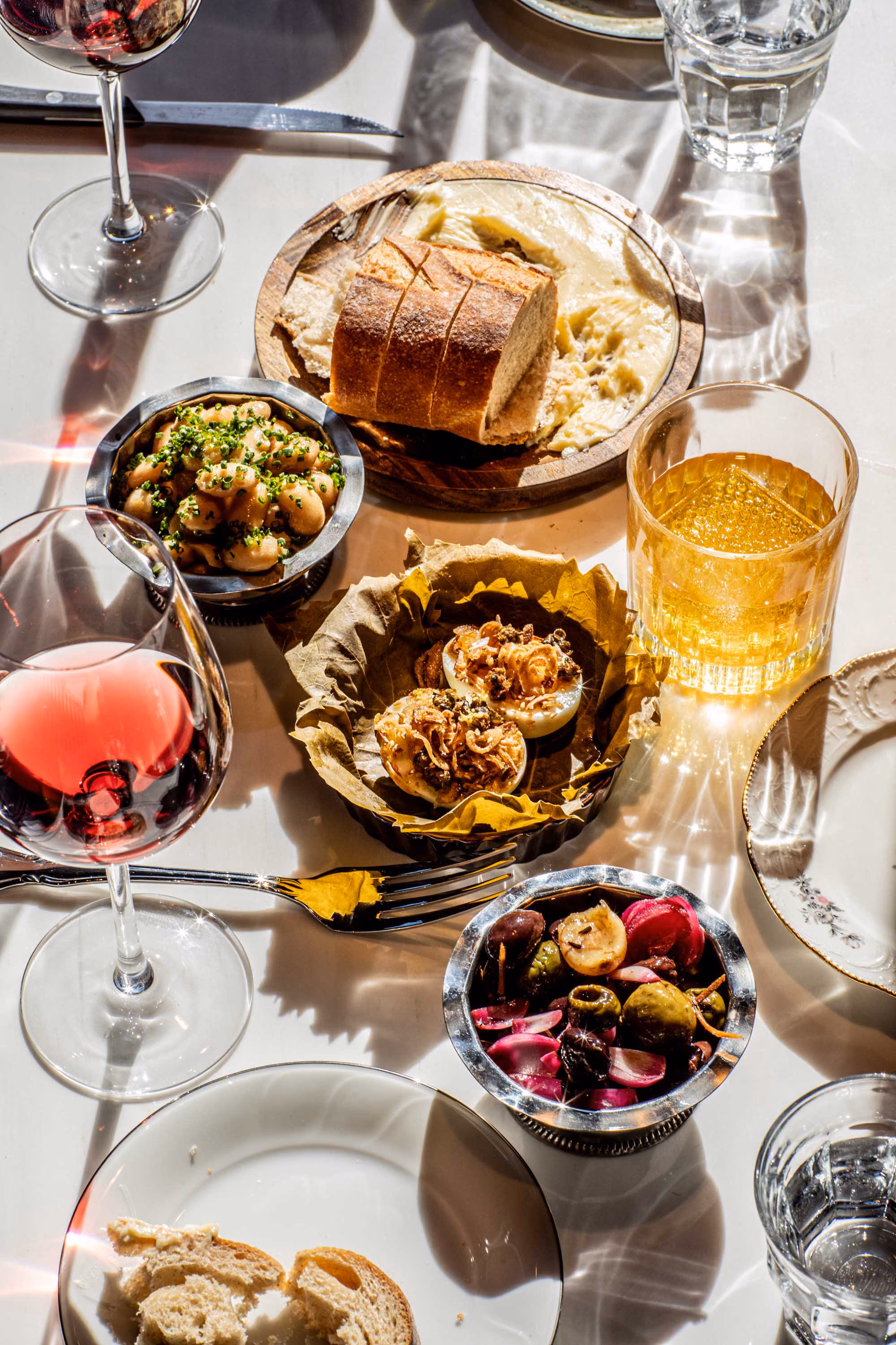 Table set with glasses of red wine and whiskey, bowls of marinated olives and beans, sliced bread on a wooden plate with butter, and deviled eggs topped with fried onions.