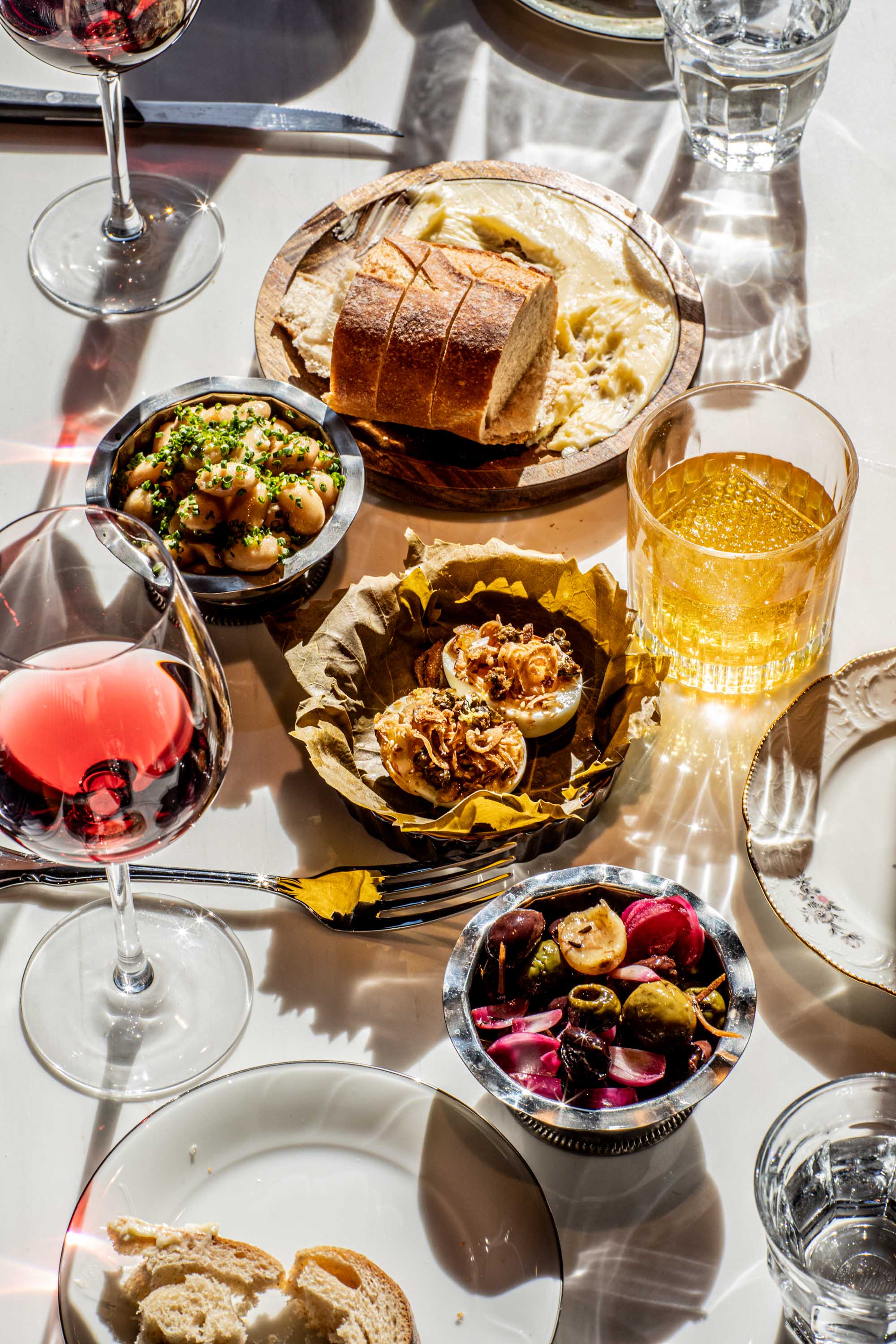 Table set with glasses of red wine and whiskey, bowls of marinated olives and beans, sliced bread on a wooden plate with butter, and deviled eggs topped with fried onions.