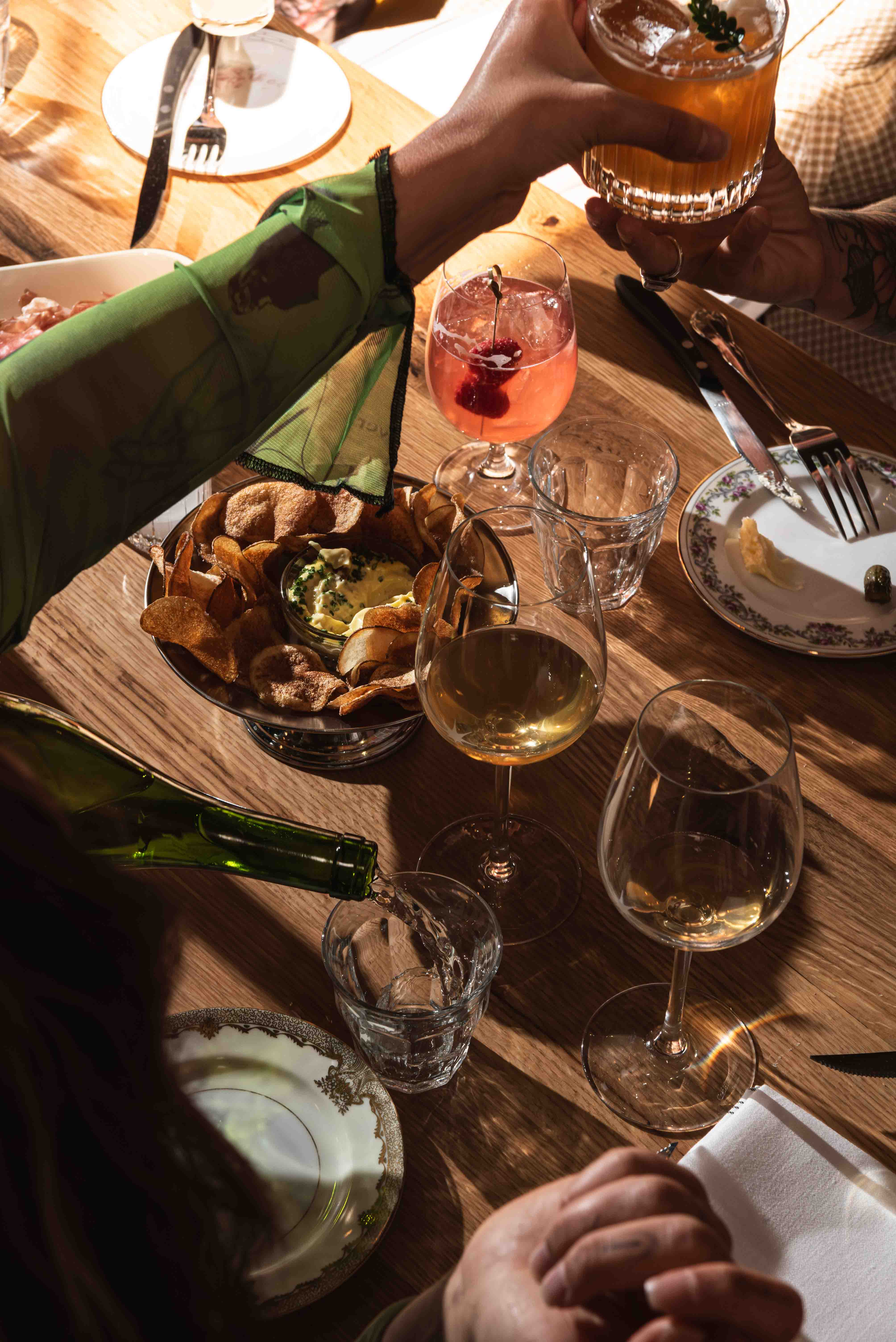 People enjoying drinks and snacks at a wooden table, with hands holding glasses, pouring wine, and a bowl of potato chips with dip in the center.