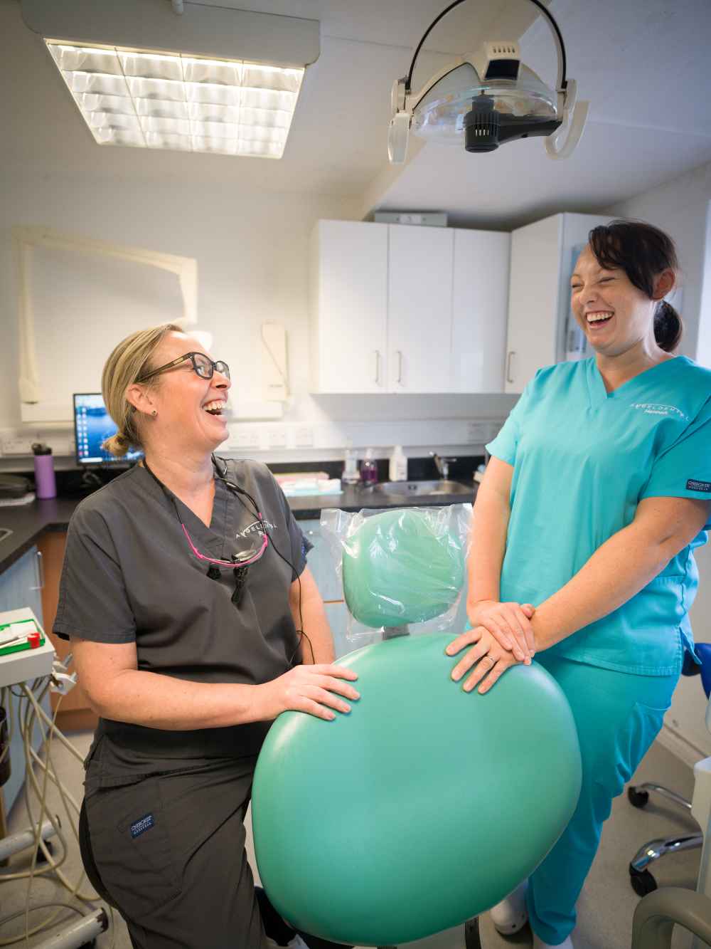 Two female dental professionals laughing together in a brightly lit dental office beside a green dental chair.
