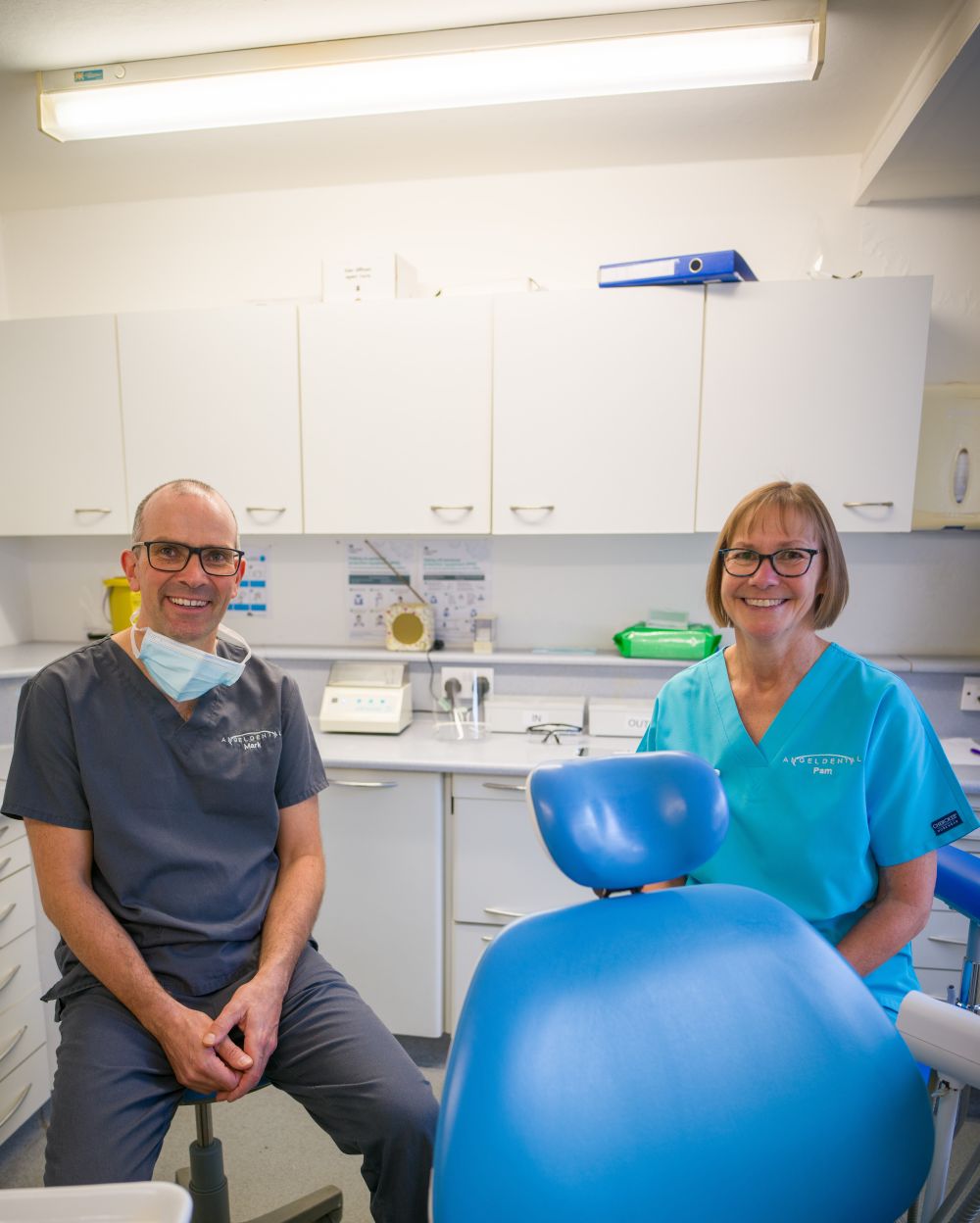 Two dental professionals smiling in a dental clinic, one wearing gray scrubs and the other in blue scrubs, with a blue dental chair in the foreground.