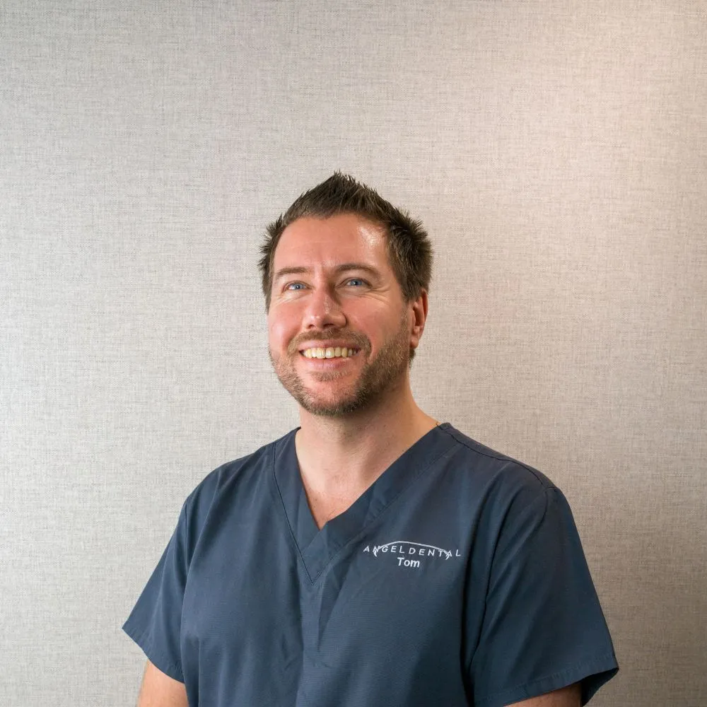 Smiling man with short spiked hair and beard wearing navy blue scrubs with Angel Dental and Tom embroidered.