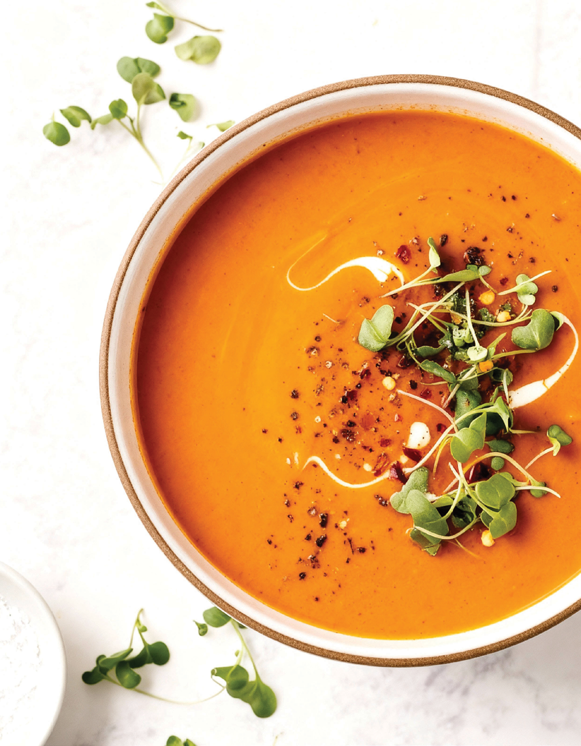 Kom met oranje pompoensoep gegarneerd met groene microgroenten en zwarte peper.