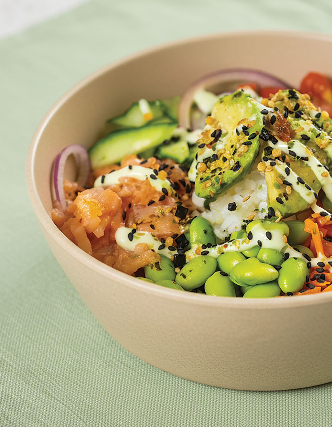 Kom met rijst, avocado, edamame, rode ui, komkommer, en stukjes rauwe zalm met sesamzaad en saus.