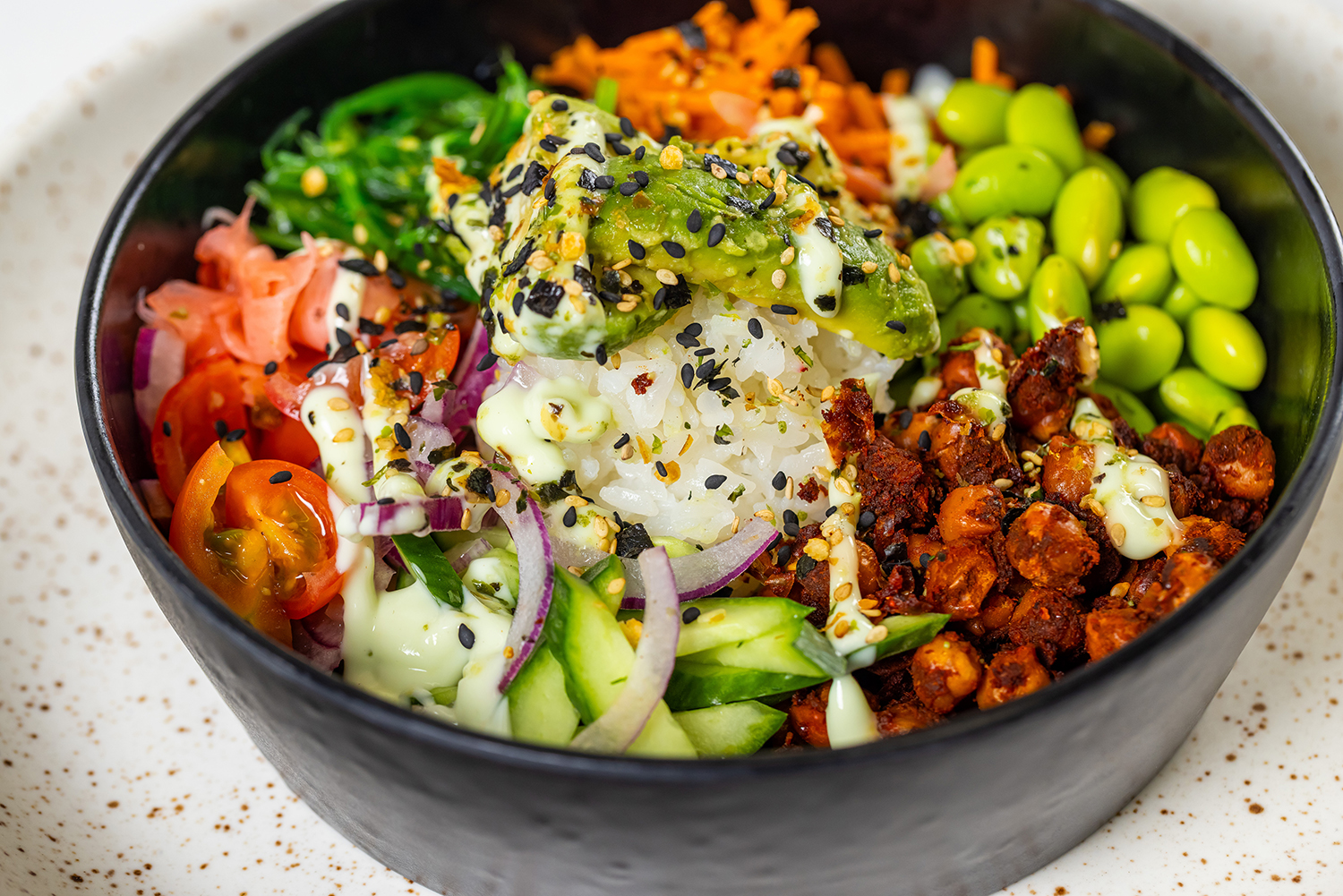 Een kleurrijke pokébowl met witte rijst, avocado, edamame, cherrytomaatjes, komkommer, wortel, rode ui, zeewiersalade en gekruide kikkererwten.