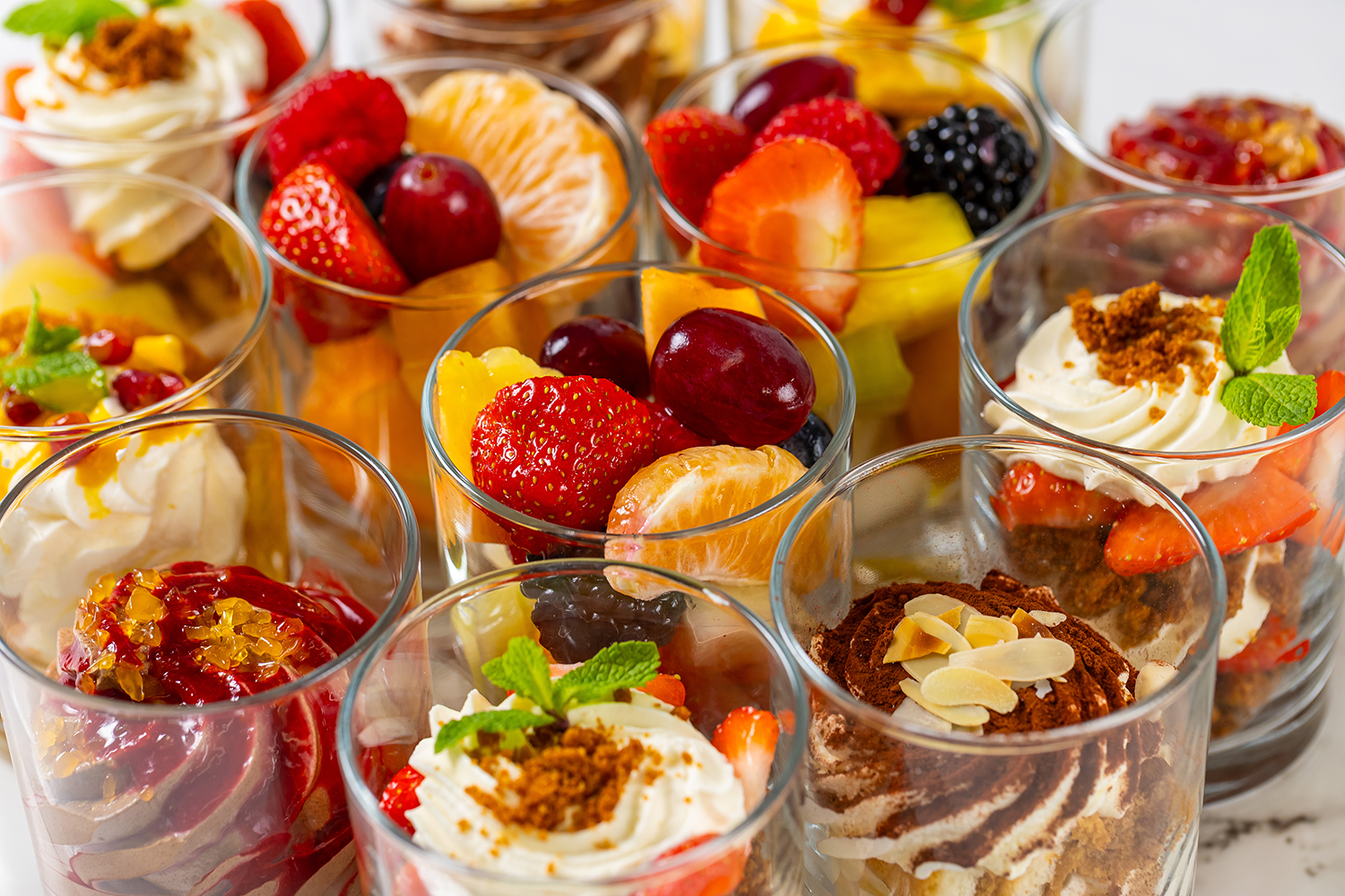 Diverse glazen gevuld met kleurrijke fruitsalades en desserts met slagroom, aardbeien, druiven en muntblaadjes.