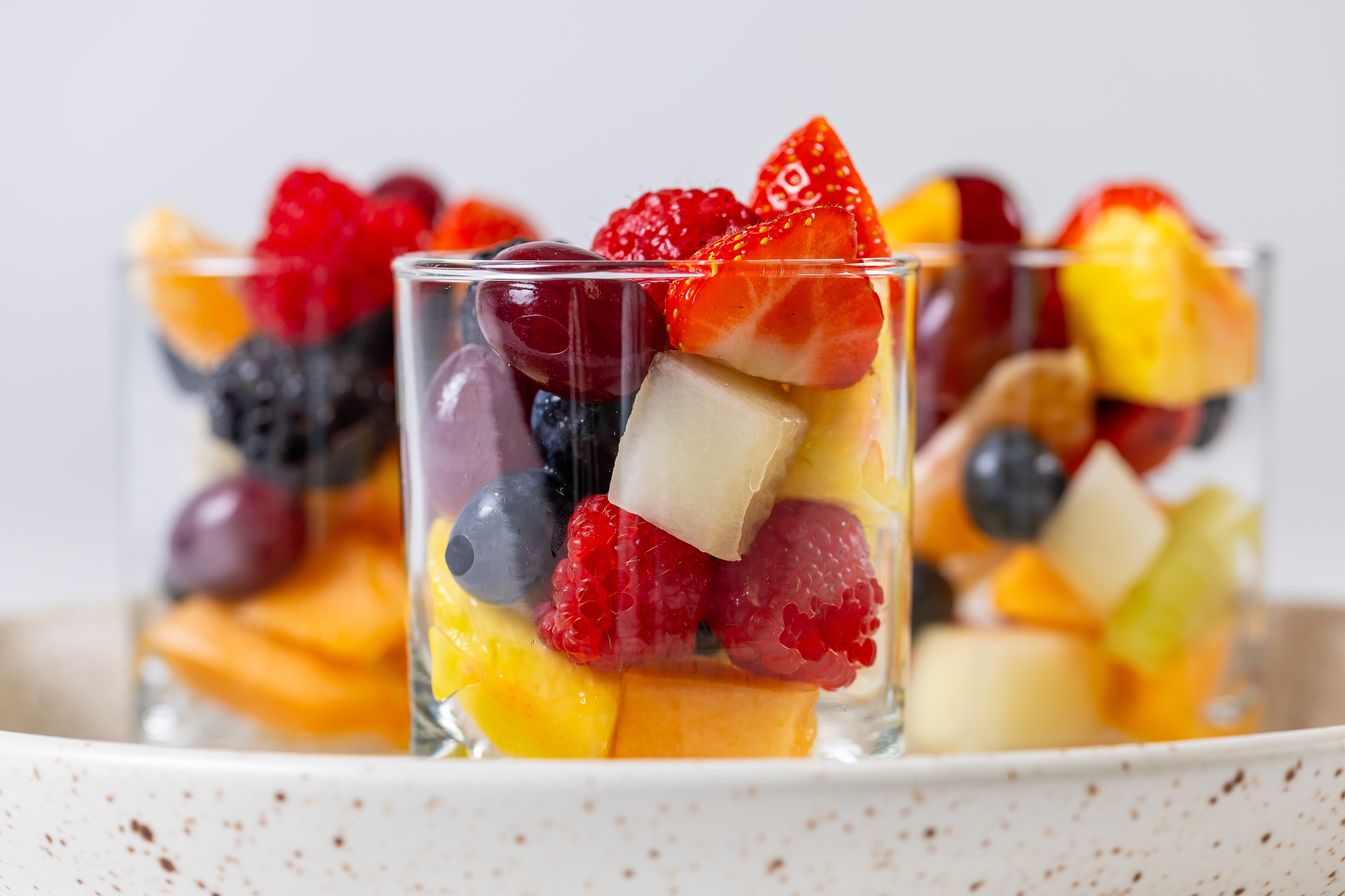 Drie glazen met diverse vers gesneden fruitsalade, waaronder aardbeien, blauwe bessen, frambozen, druiven en meloen.