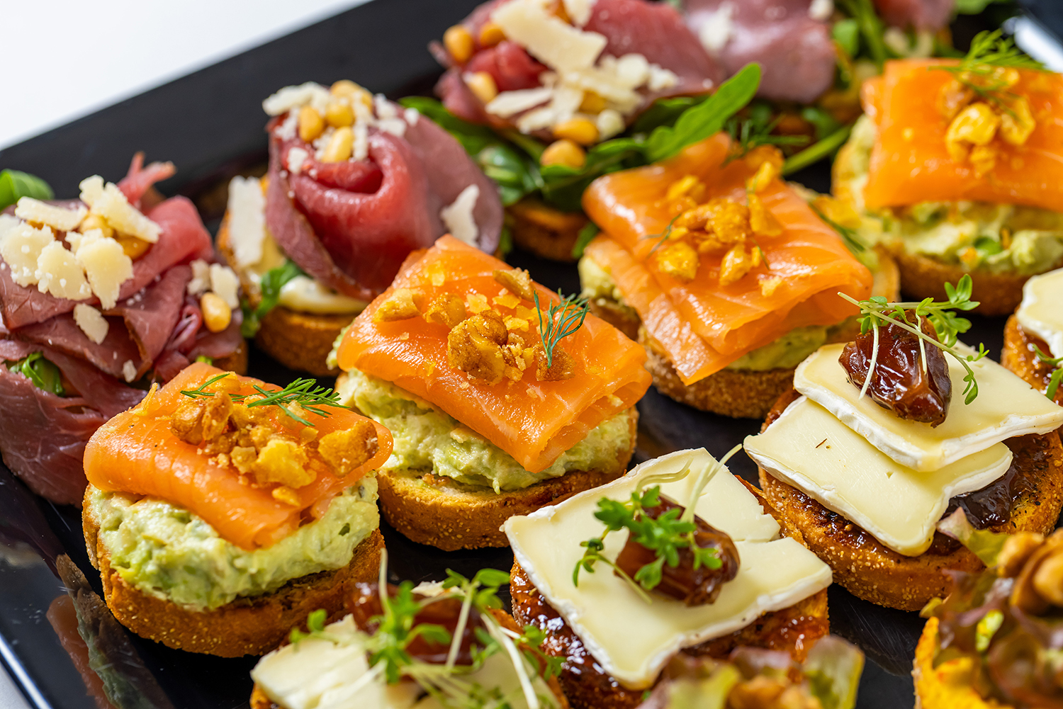 Een zwart dienblad met diverse luxe hapjes, waaronder toastjes met gerookte zalm en dille, toastjes met kaas en dadels, en toastjes met rosé vlees en kaas.
