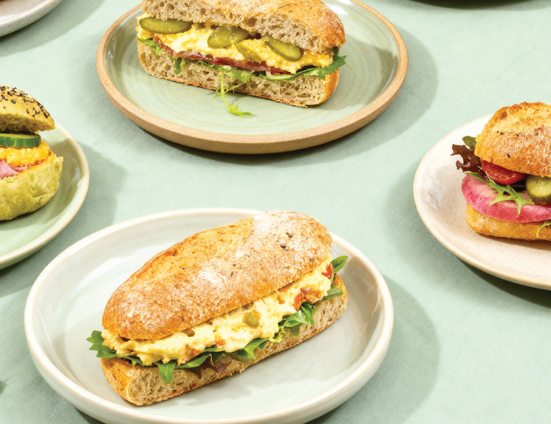 Vier verschillende sandwiches met eier- en groentenvulling op lichte borden, geplaatst op een lichtgroene tafel.