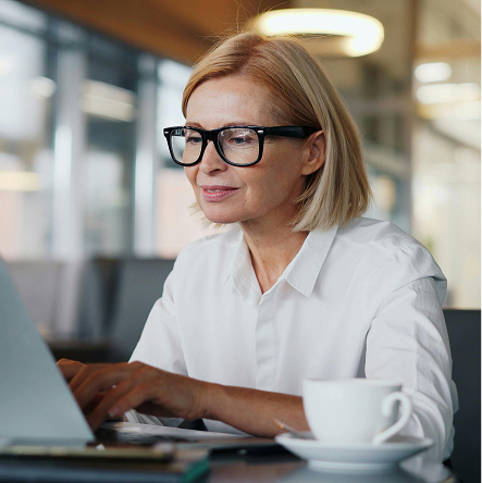 Middle-aged woman wearing glasses and a white shirt working on a laptop with a coffee cup nearby in an office setting.