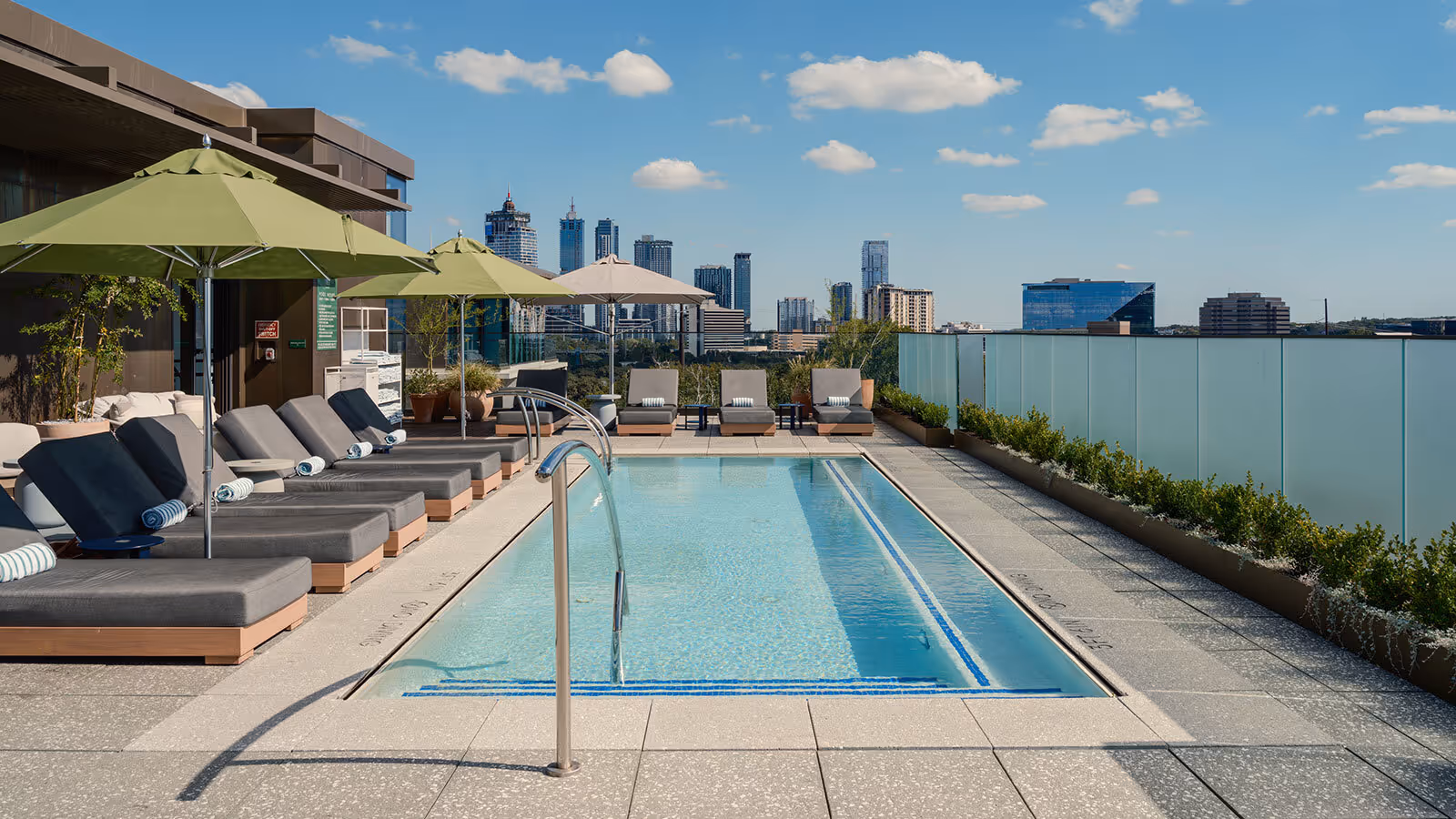 Rooftop pool with lounge chairs, green umbrellas, and city skyline in the background under a partly cloudy sky.