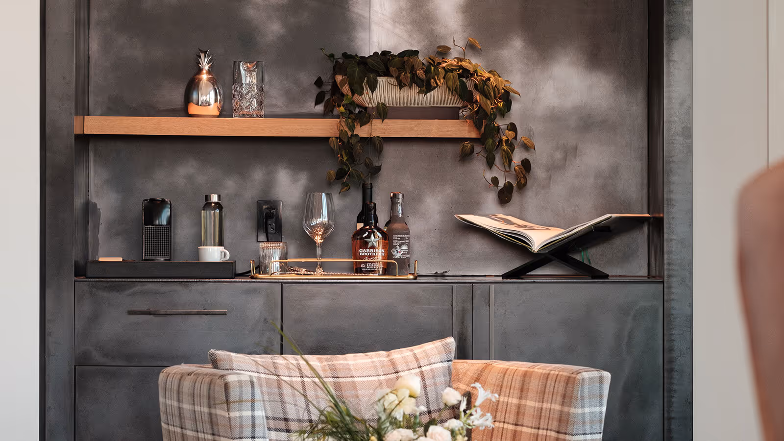 Modern home bar setup with bottles, glasses, a coffee machine, and a plant on wooden shelves above a plaid armchair.